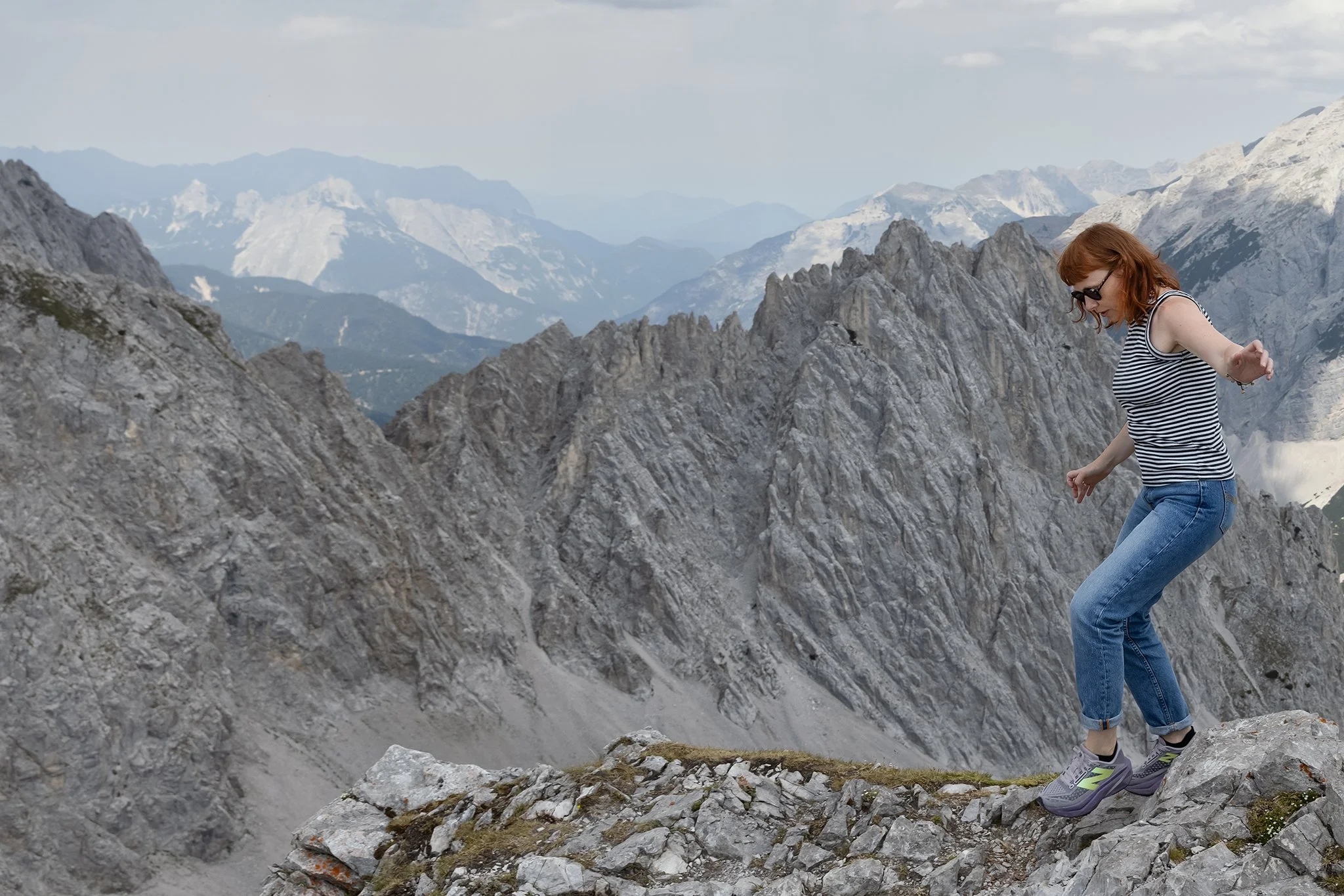 A woman with red hair, sunglasses, wearing a striped tank top, blue jeans, and sneakers, balancing on rocky mountain terrain with a snowy mountain range in the background.