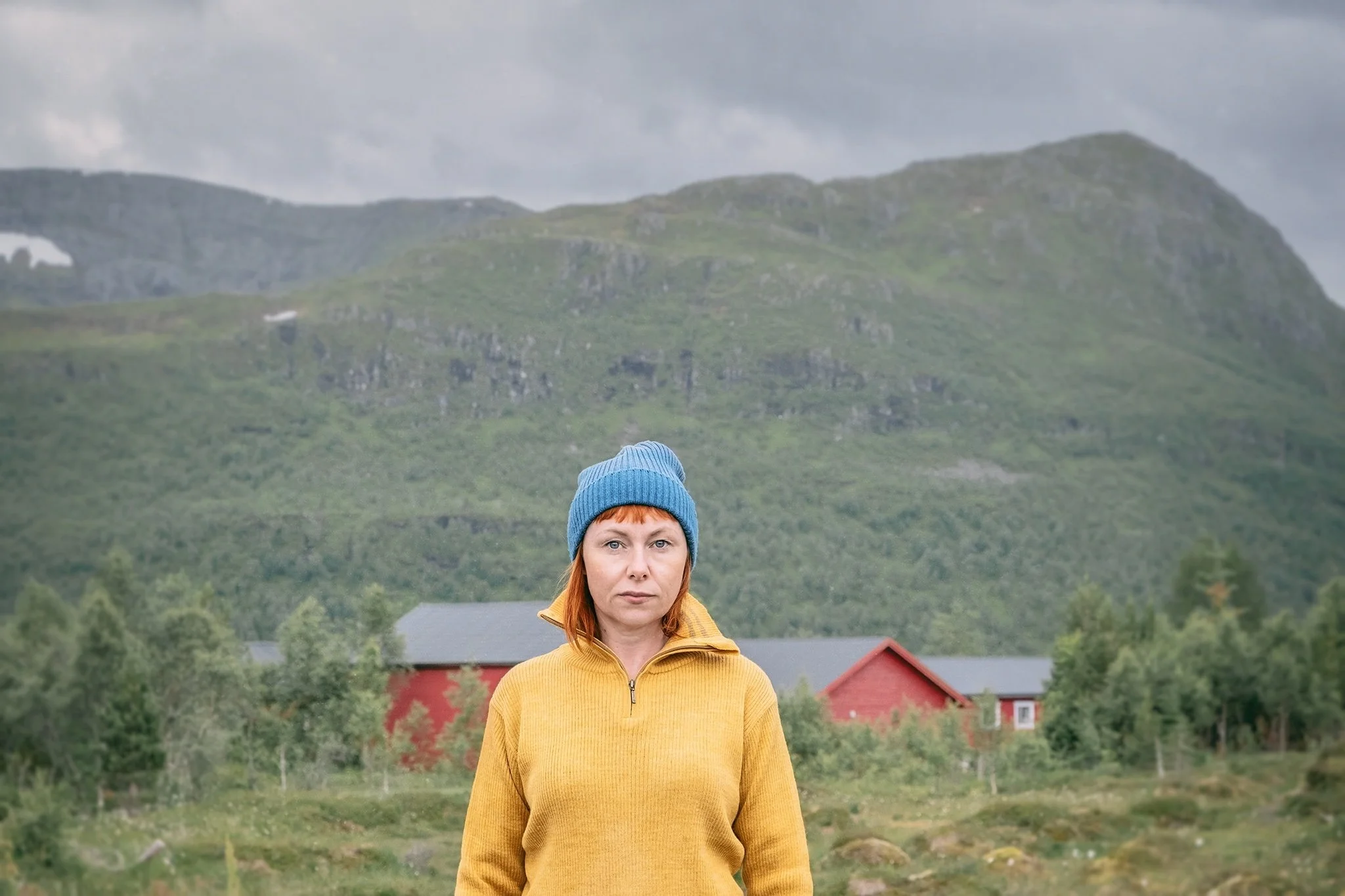 A woman with red hair wearing a blue beanie and yellow jacket standing outdoors in front of green hills and a red barn.