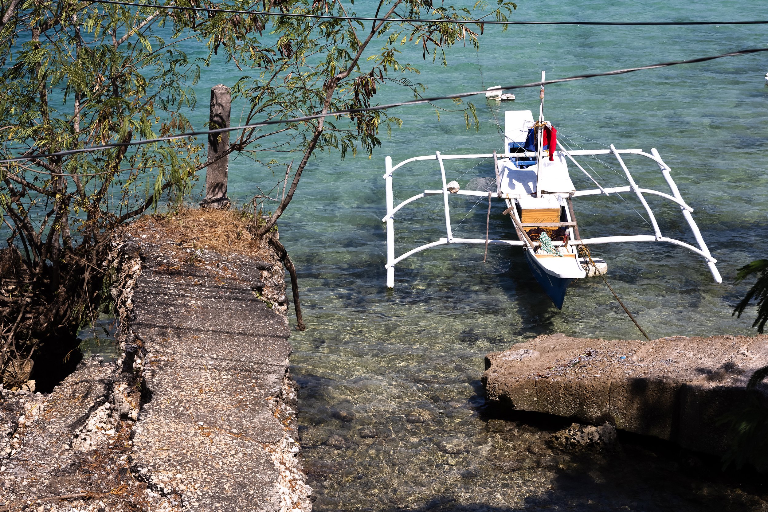 A small boat with a white frame is anchored near a rocky shoreline with clear greenish water. There is a tree with green leaves partially covering the view.