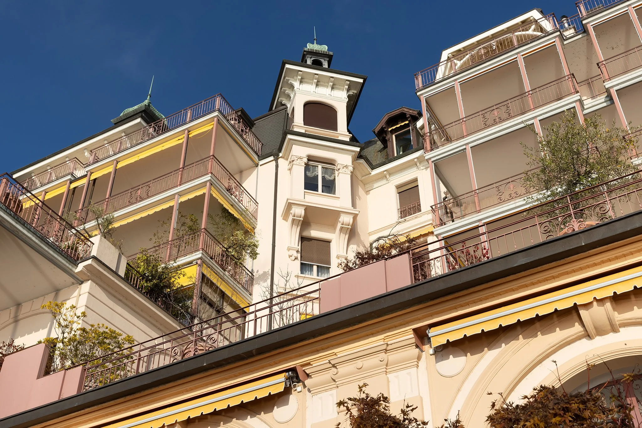 Lower view of a multi-story building with balconies, decorative railings, and a tower with a clock and spire against a clear blue sky.