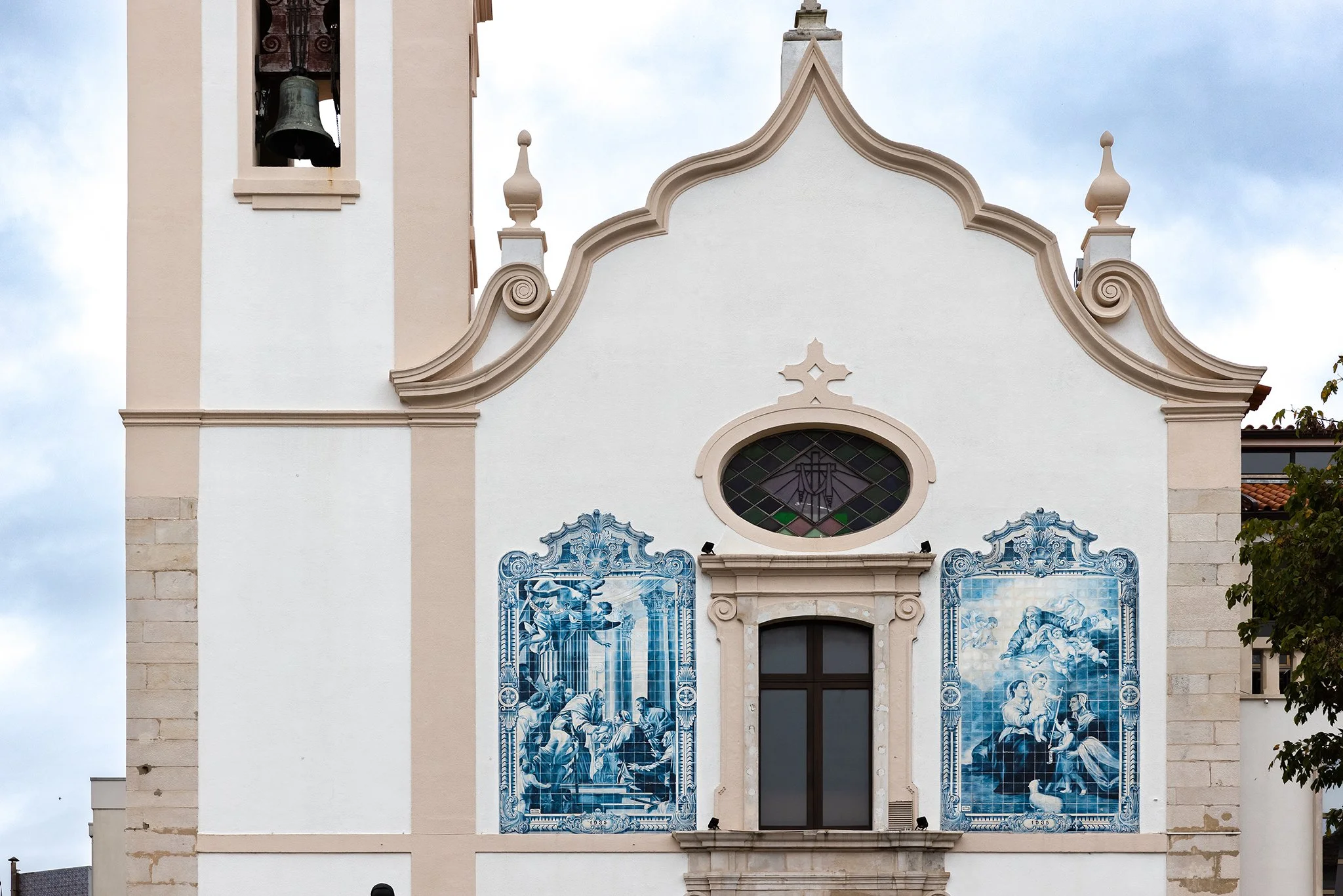 The facade of a historic building featuring a bell tower on the left and blue and white azulejo tile murals depicting religious scenes, topped with ornate architectural details.