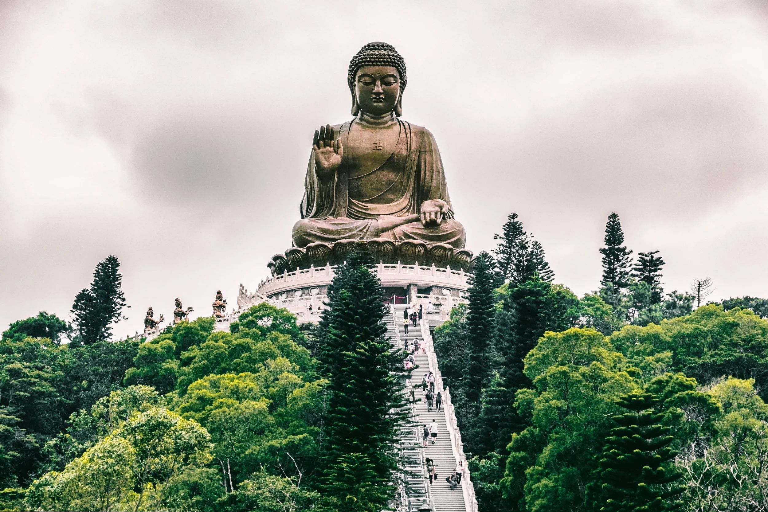 Large bronze statue of Buddha sitting on a pedestal with a stairway leading up to it, surrounded by lush green trees and overcast sky.
