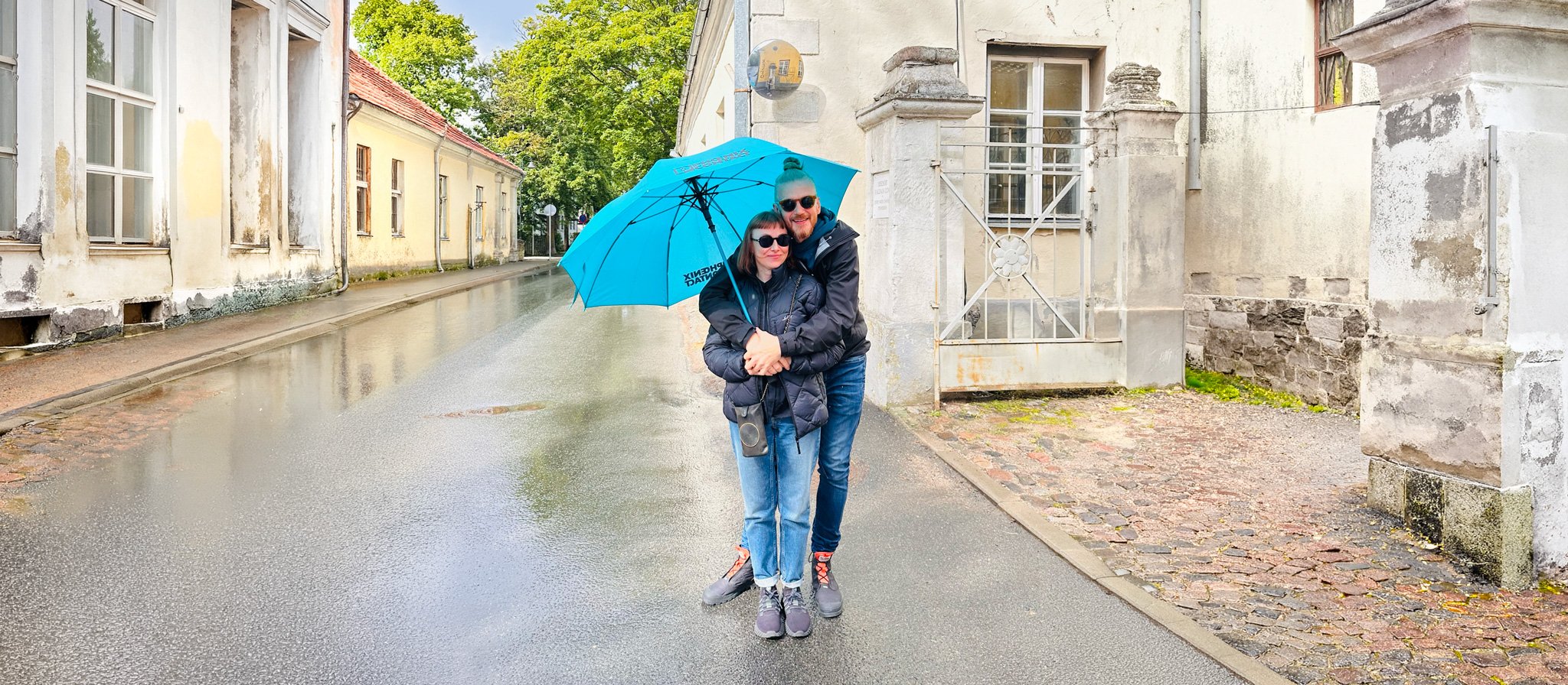 A couple standing under a blue umbrella in the rain on a wet cobblestone street, surrounded by old buildings with white and gray walls, and green trees in the background.