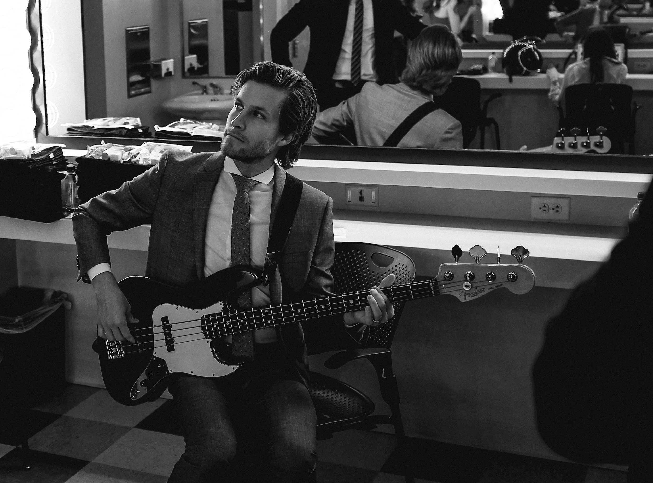 A man in a suit with a guitar sitting in a dressing room, reflected in a mirror with makeup and styling supplies around, and a woman with short hair sitting in the background.