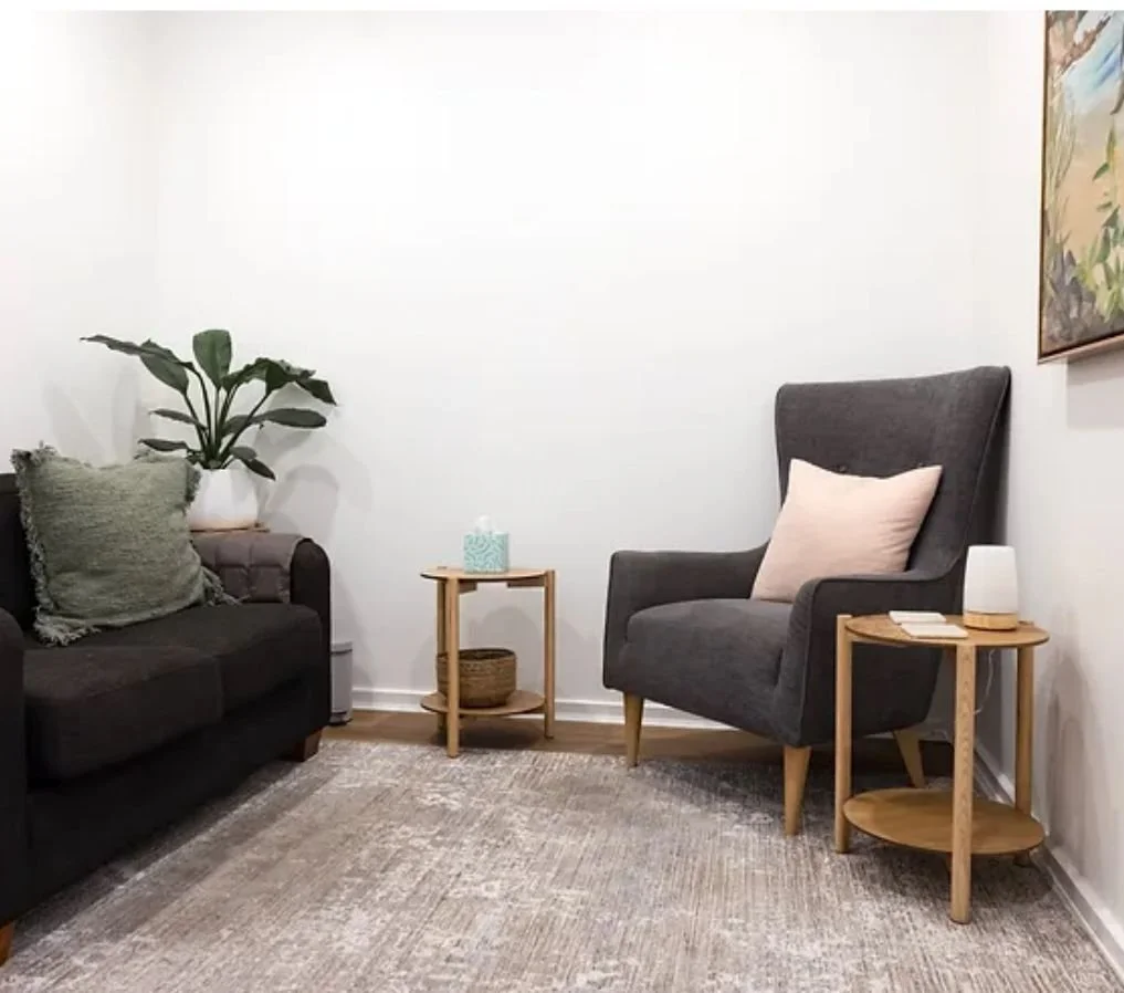 A cozy living room corner with a gray armchair and a black sofa, side tables holding a tissue box, a lamp, and a decorative item, a large plant on the sofa's armrest, and a painting on the wall.