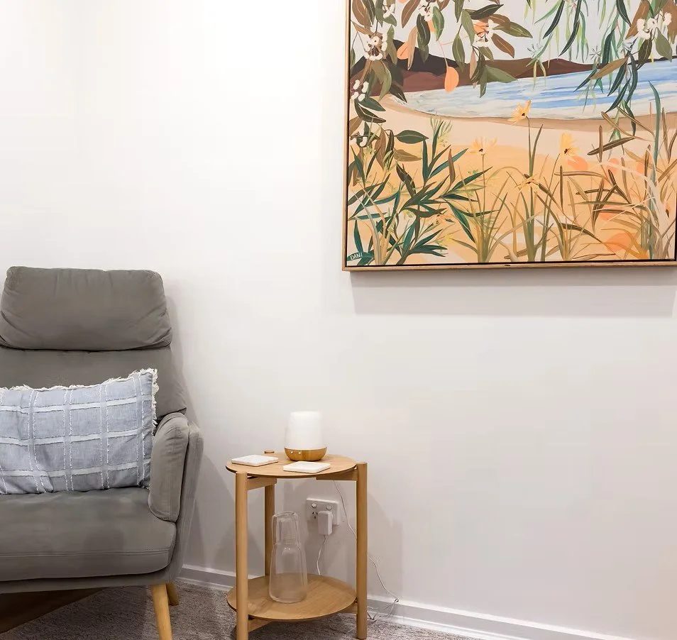 A living room corner with a gray sofa, a light wood side table holding a white lamp and a book, a glass pitcher inside the table's lower shelf, and a large painting of a beach scene hanging on the white wall.