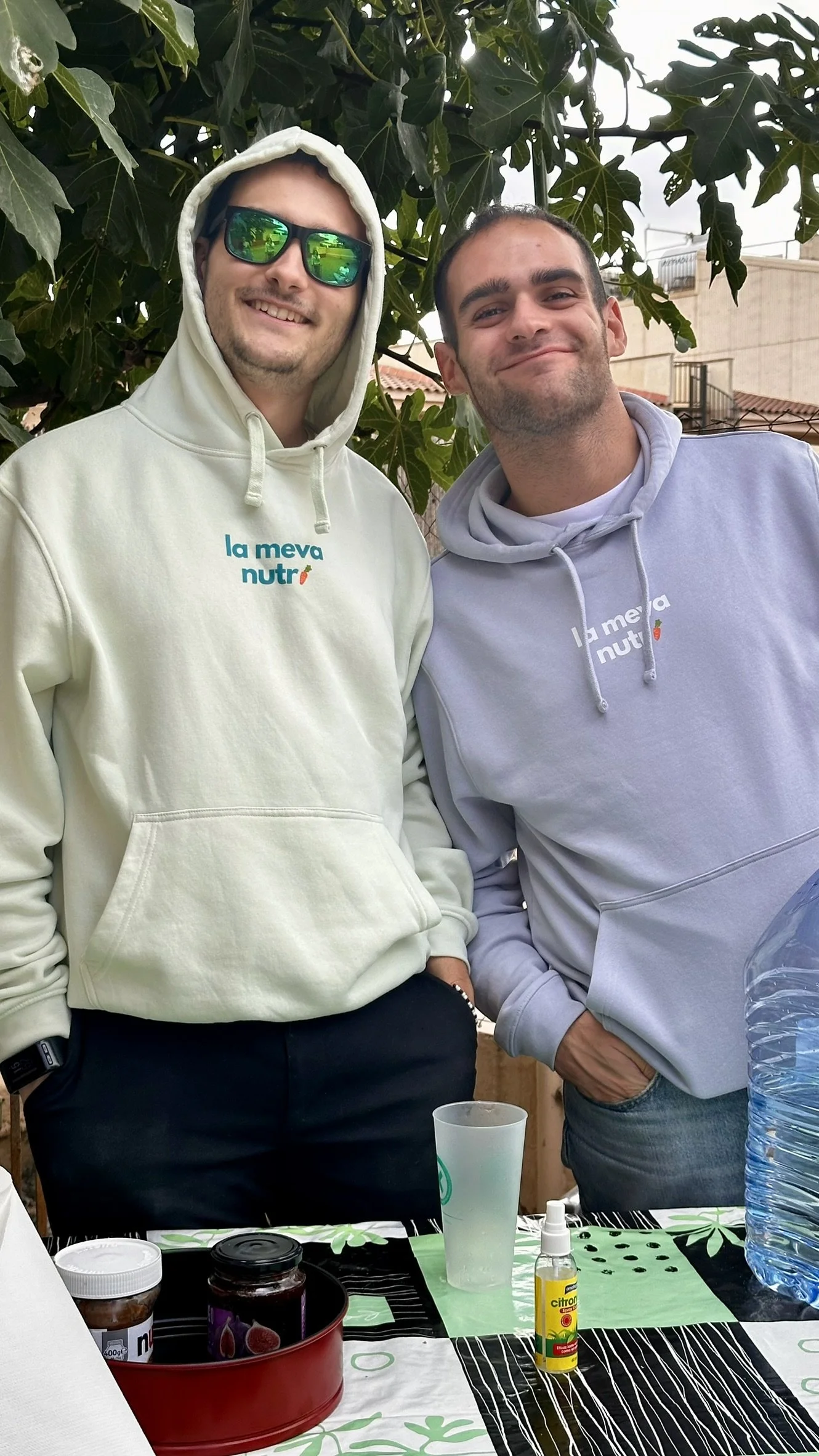 Dos hombres sonriendo, uno con gafas de sol y sudadera blanca, el otro con sudadera gris, están en un ambiente al aire libre con árboles de follaje denso. En la mesa hay varios objetos: frascos, un vaso de plástico, un repelente y una botella de agua