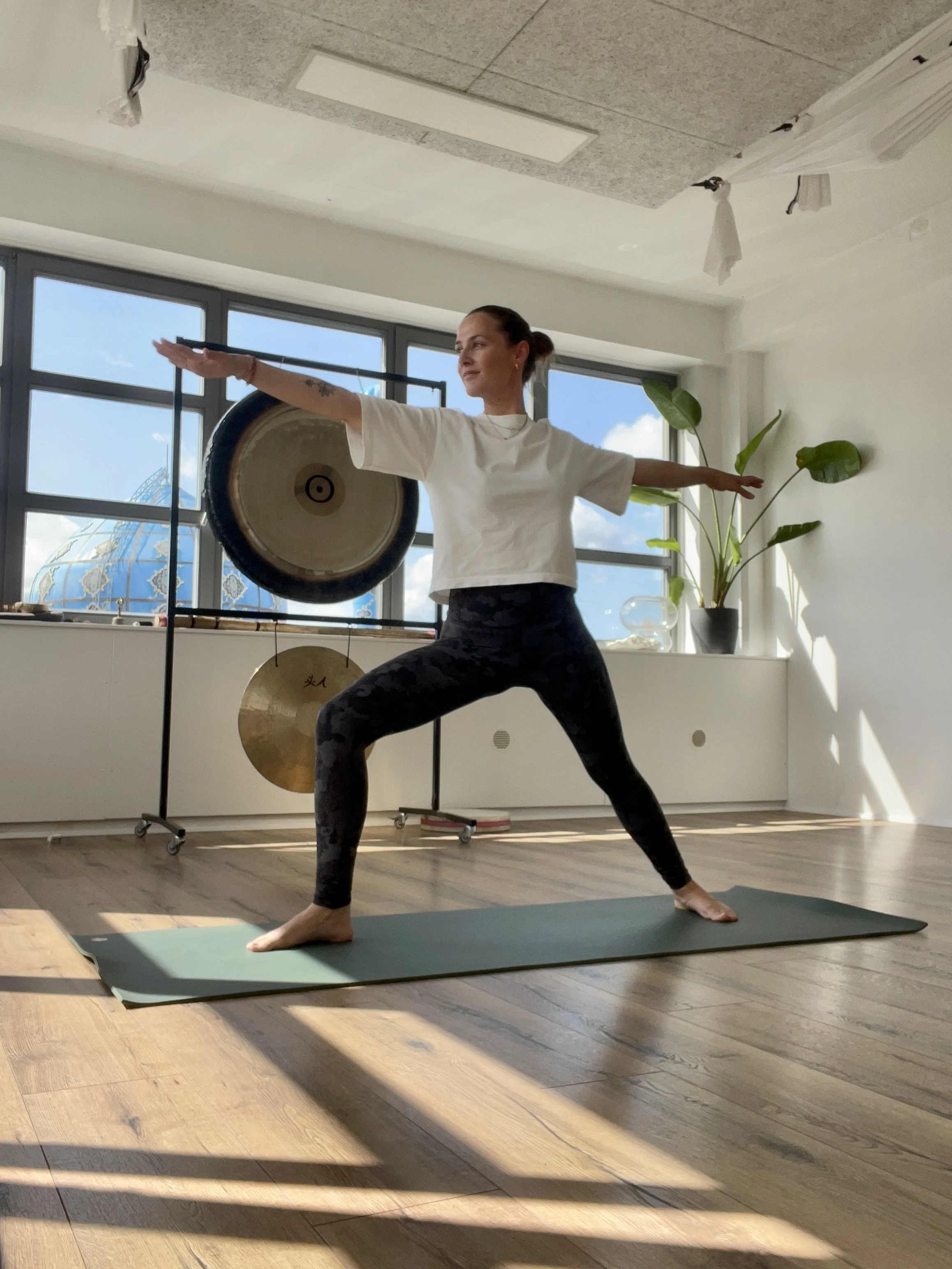Psykoterapeut Anna Nordkvist i stående yogastilling på yogamåtte i et lyst rum med store vinduer og planter.