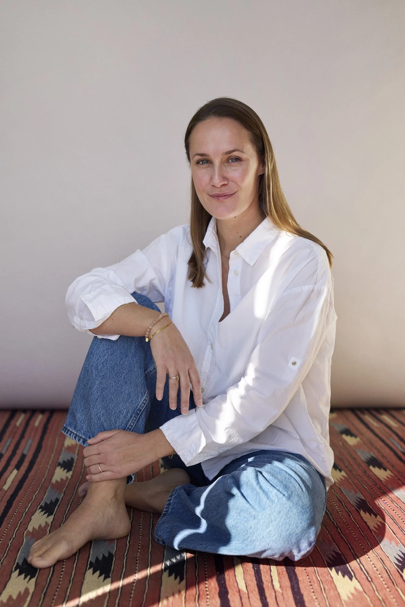 Psykoterapeut Anna Nordkvist sidder afslappet på gulvet, iført en hvid skjorte og blå jeans, med et roligt smil, foran en lys væg og et farverigt tæppe.