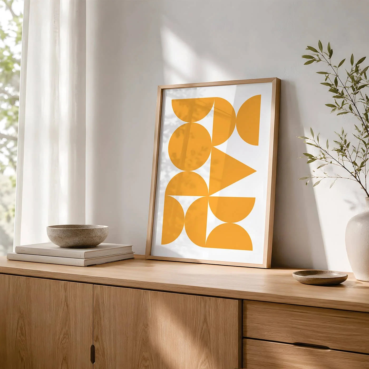 Yellow Abstract Forms Print presented in a wooden frame on a contemporary wooden sideboard with books, a decorative bowl and a houseplant by a window.