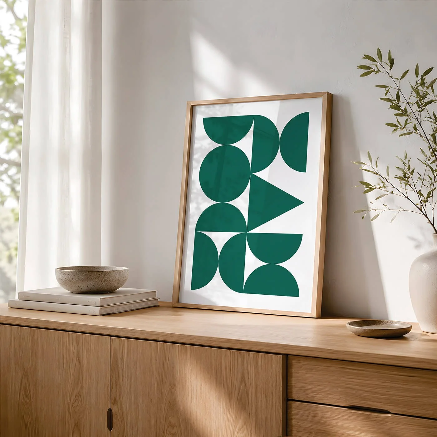 Green Abstract Forms Print styled in a wooden frame on a contemporary sideboard with books, a decorative bowl and a houseplant beside a window.