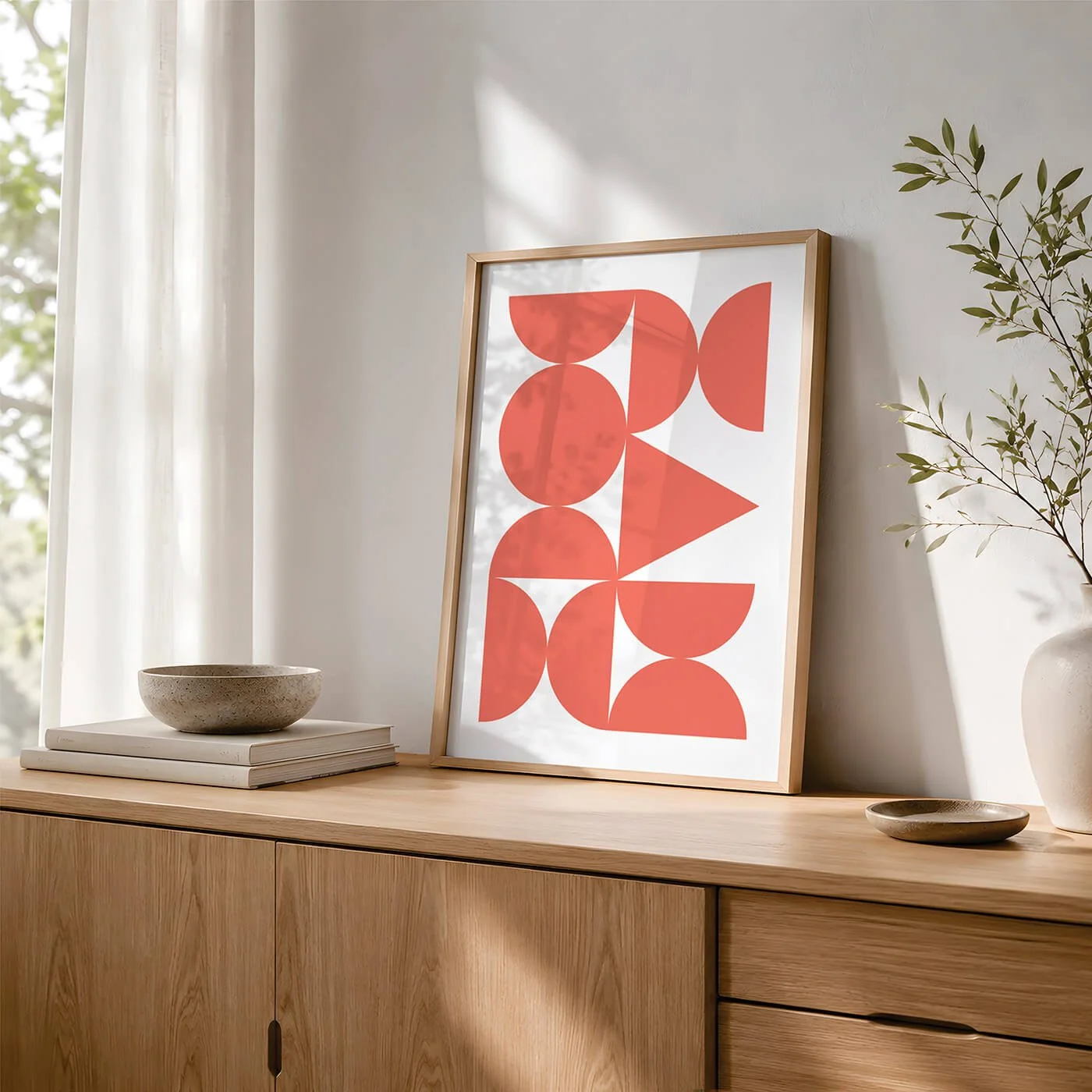 Coral Abstract Forms Print in a wooden frame styled on a contemporary sideboard with decorative books, a modern bowl and a plant next to a window.