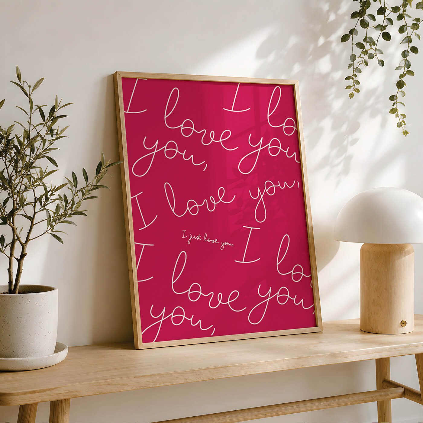 A pink framed wall art with white cursive writing saying 'I love you' repeatedly in a circle pattern, with smaller text in the center saying 'I just love you', positioned on a wooden table next to a potted plant and a modern lamp.