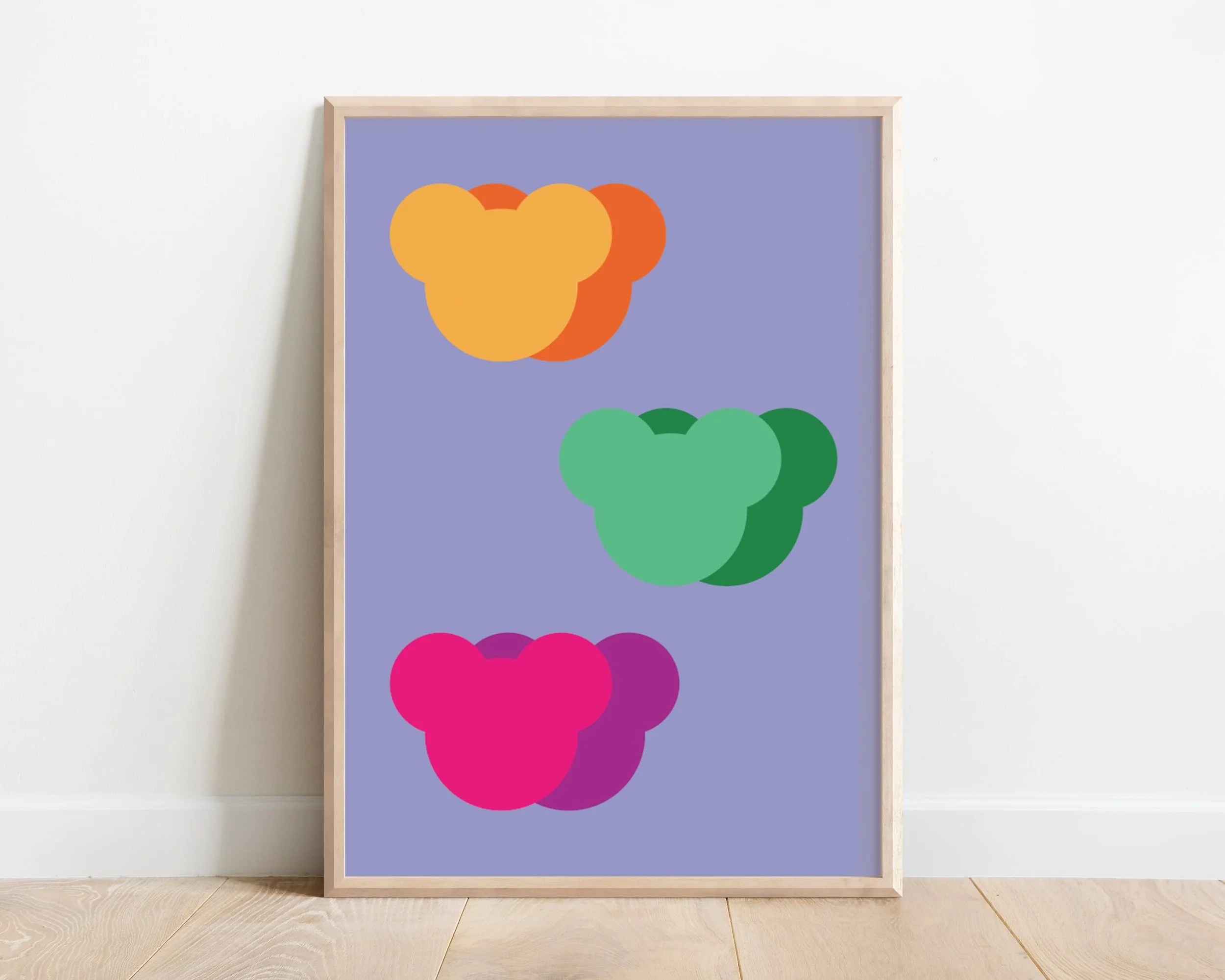 Framed abstract teddy bear heads print in pink, green, and yellow on a purple background, styled against a plain wall and wooden floor.