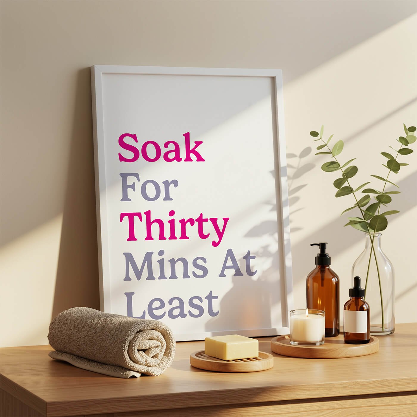 A contemporary pastel bathroom typography print featuring lilac and hot pink text reading Soak For Thirty Mins At Least, styled in a wooden frame styled next to a stack of towels