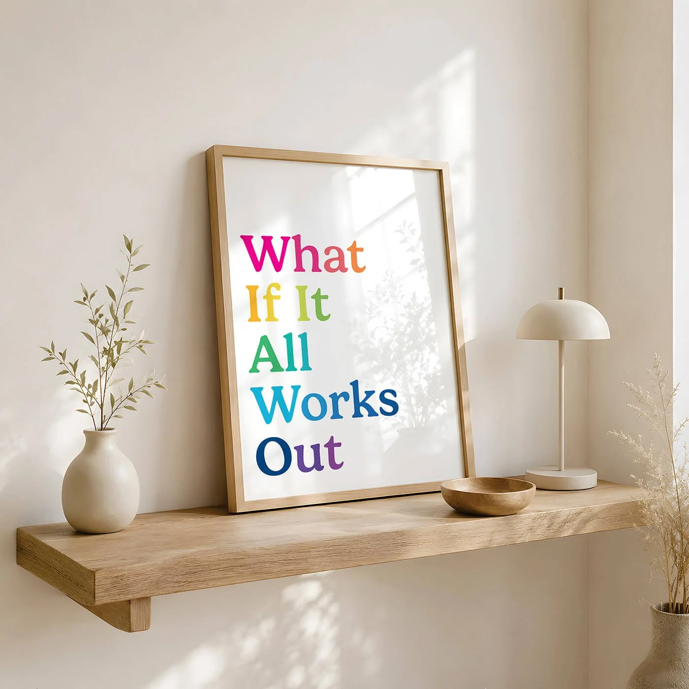 A mid century rainbow typography print reading What If It All Works Out, displayed in a wooden frame on a natural timber floating shelf with a neutral ceramic vase and a small wooden bowl