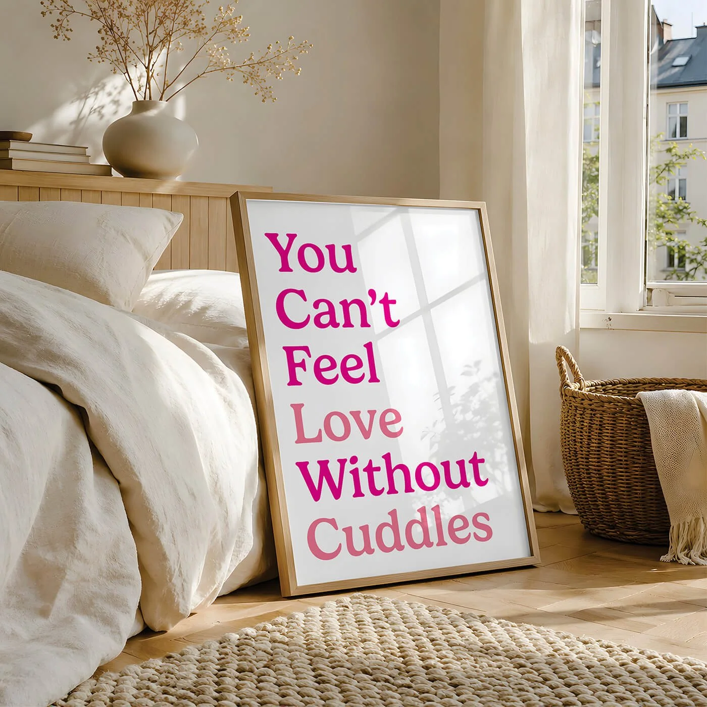 A framed quote poster leaning against a bed reads, "You Can't Feel Love Without Cuddles" in pink and light pink text, with a bright sunny window in the background.