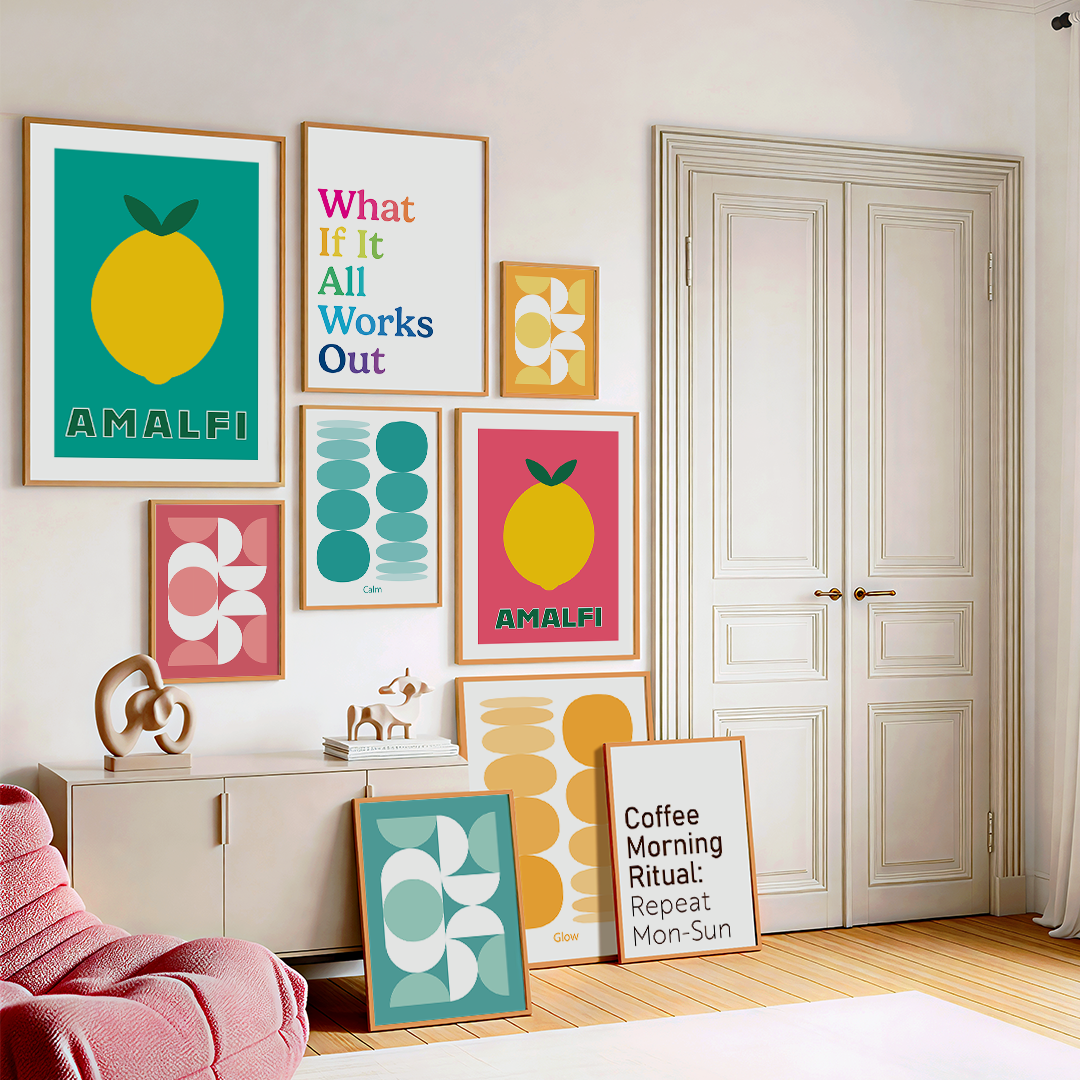 A room with a white wall decorated with colorful framed posters featuring modern graphic art and positive messages. The posters include images of lemons, abstract shapes, and text such as "What If It All Works Out" and "AMALFI." There is a white cabinet with sculptural decor on top, a pink textured armchair, and a wooden floor with a white curtain on the right side.