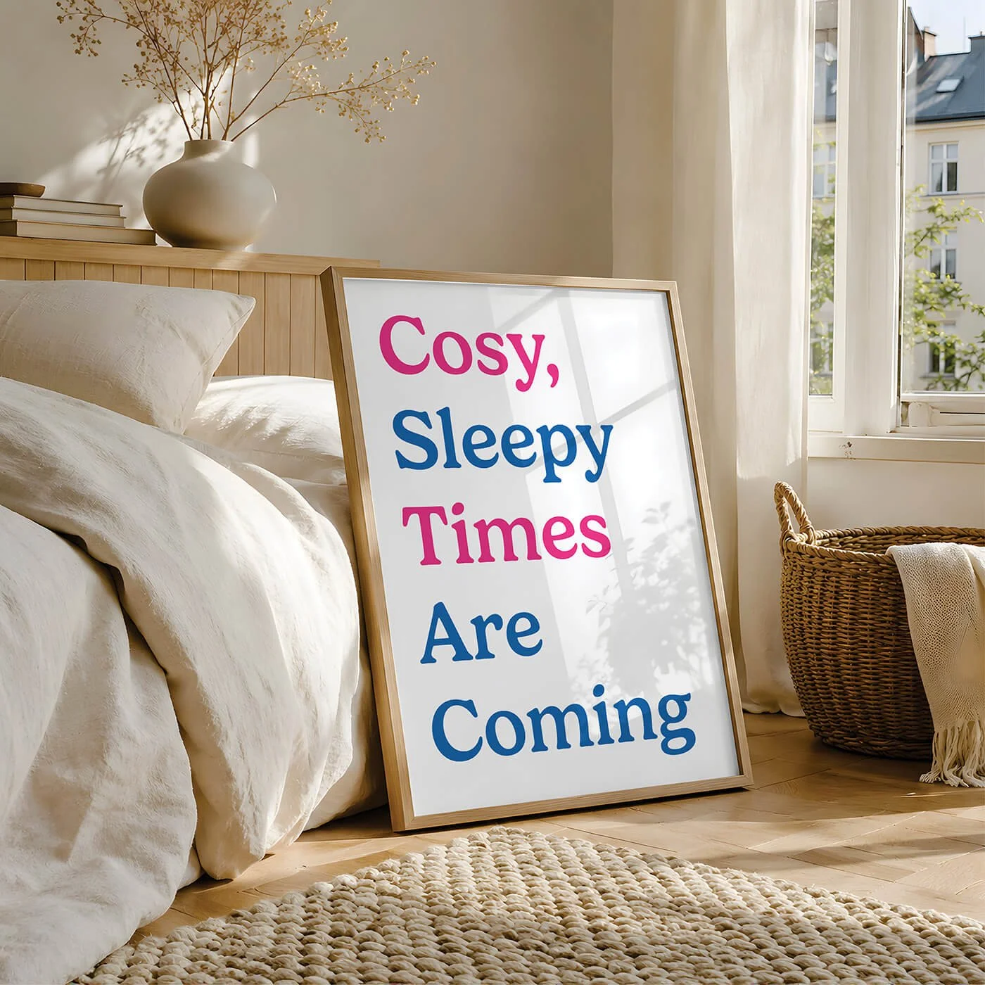 A cool bedroom typography art print with blue and pink lettering reading Cosy Sleepy Times Are Coming, in a wooden frame leaning against a light wood bed frame by a sunny window