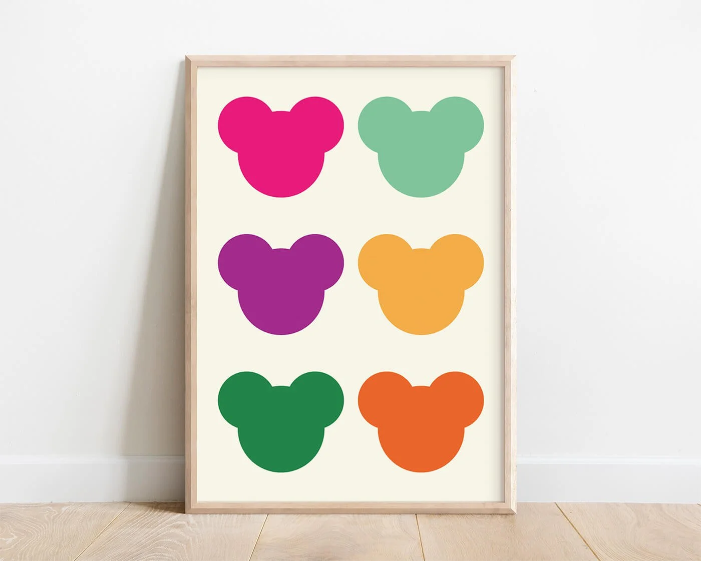 Framed abstract teddy bear heads print in pink, green, and yellow on a white background, styled against a plain wall and wooden floor.