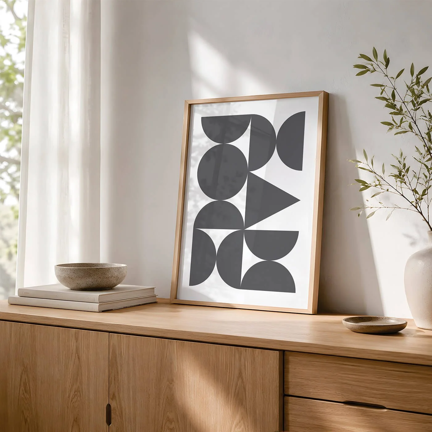 Grey Abstract Forms Print displayed in a wooden frame on a modern sideboard with stacked books, a ceramic bowl, a leafy plant and natural window light.