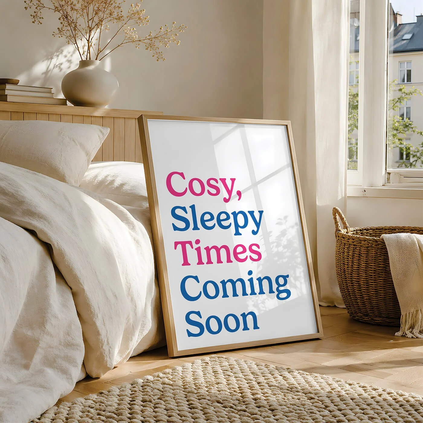 A cool bedroom typography art print with blue and pink lettering reading Cosy Sleepy Times Are Coming, in a wooden frame leaning against a light wood bed frame by a sunny window