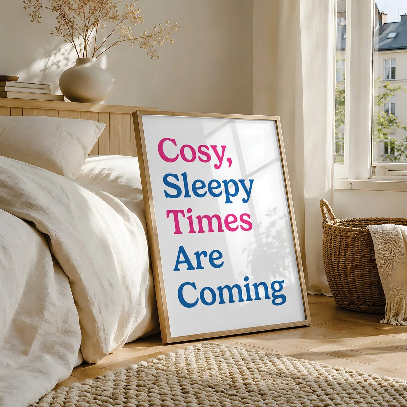 A cool bedroom typography art print with blue and pink lettering reading Cosy Sleepy Times Are Coming, in a wooden frame leaning against a light wood bed frame by a sunny window