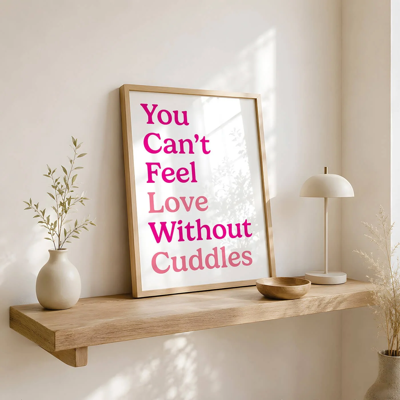 A funky pink quote typography print reading You Cant Feel Love Without Cuddles, styled in a light wooden frame resting on a natural wood floating shelf next to a modern cream table lamp