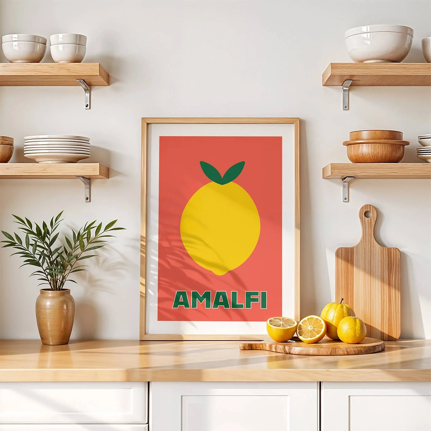 A framed terracotta orange Amalfi lemon art print displayed on a minimalist worktop within a contemporary white kitchen interior.