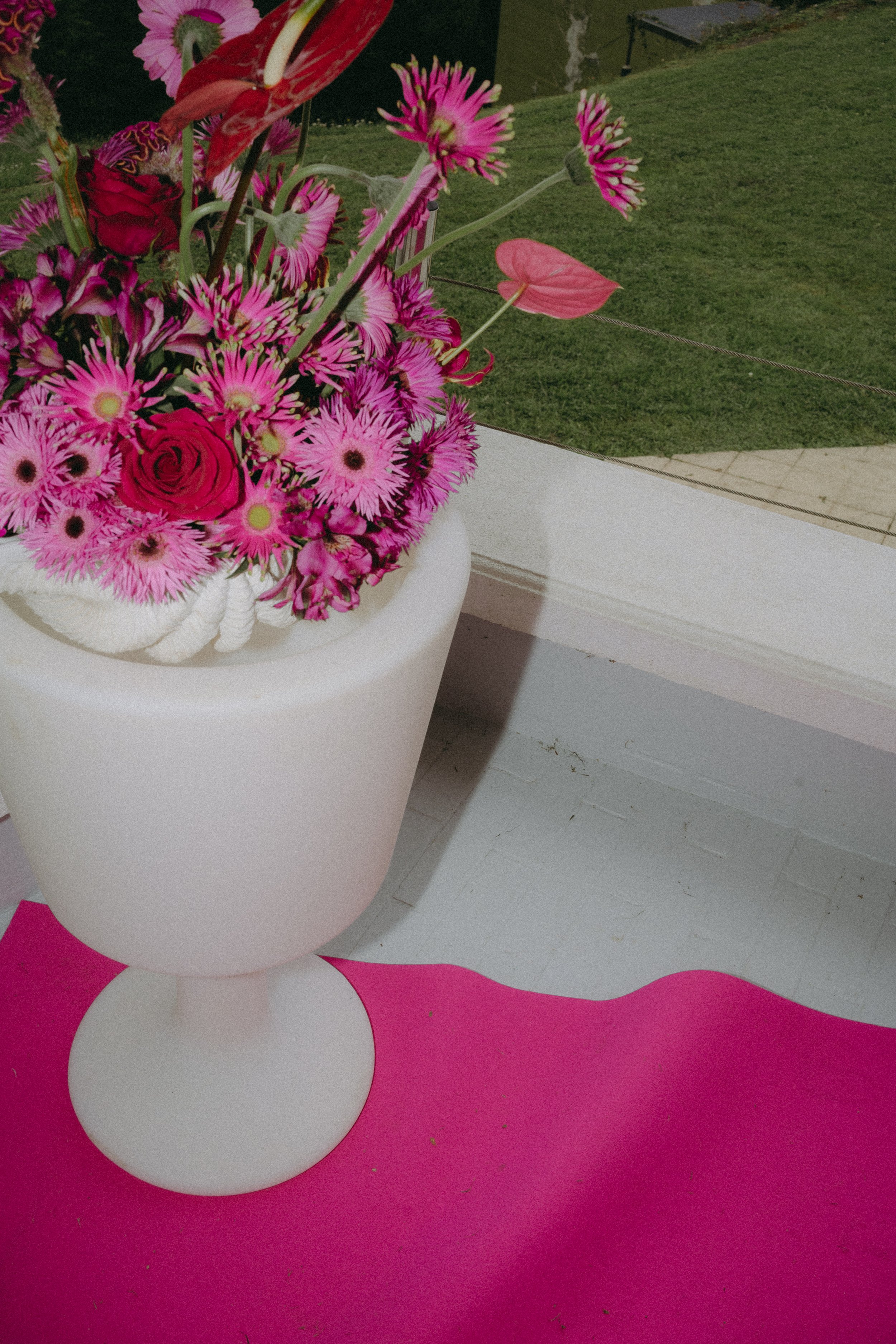 Un vase blanc avec des fleurs roses et rouges, sur une surface rose, devant une pelouse verte.