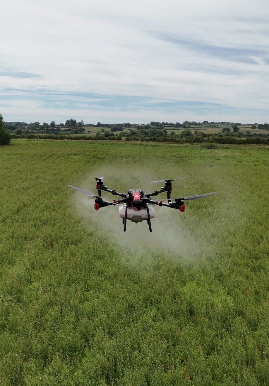 A drone flying low over a green field in a rural area with trees and rolling hills in the background under partly cloudy skies.