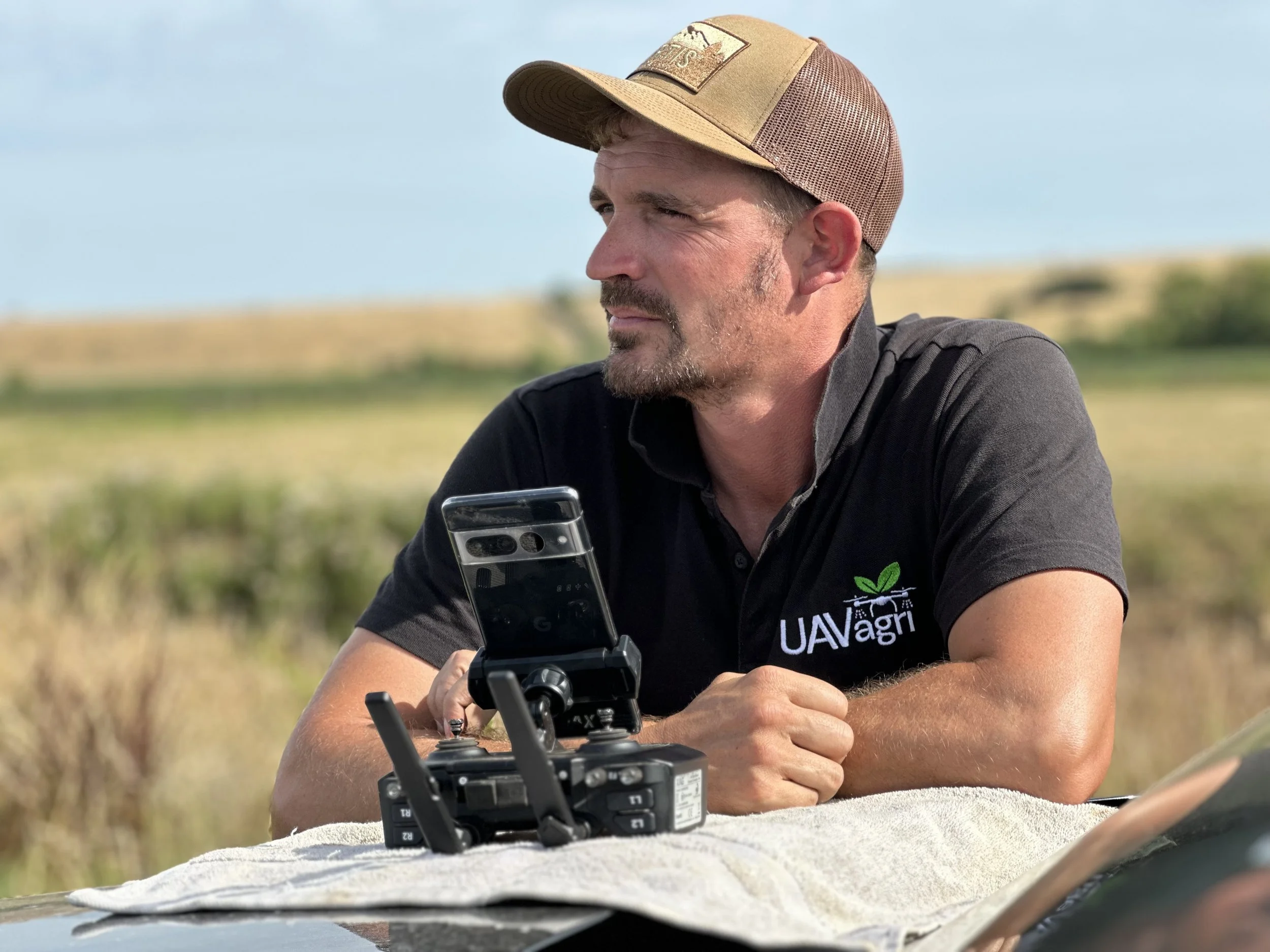 A man wearing a tan and brown baseball cap and a black T-shirt is operating a drone controller outdoors in a grassy field. He is holding a smartphone while looking at it, with a clear blue sky in the background.