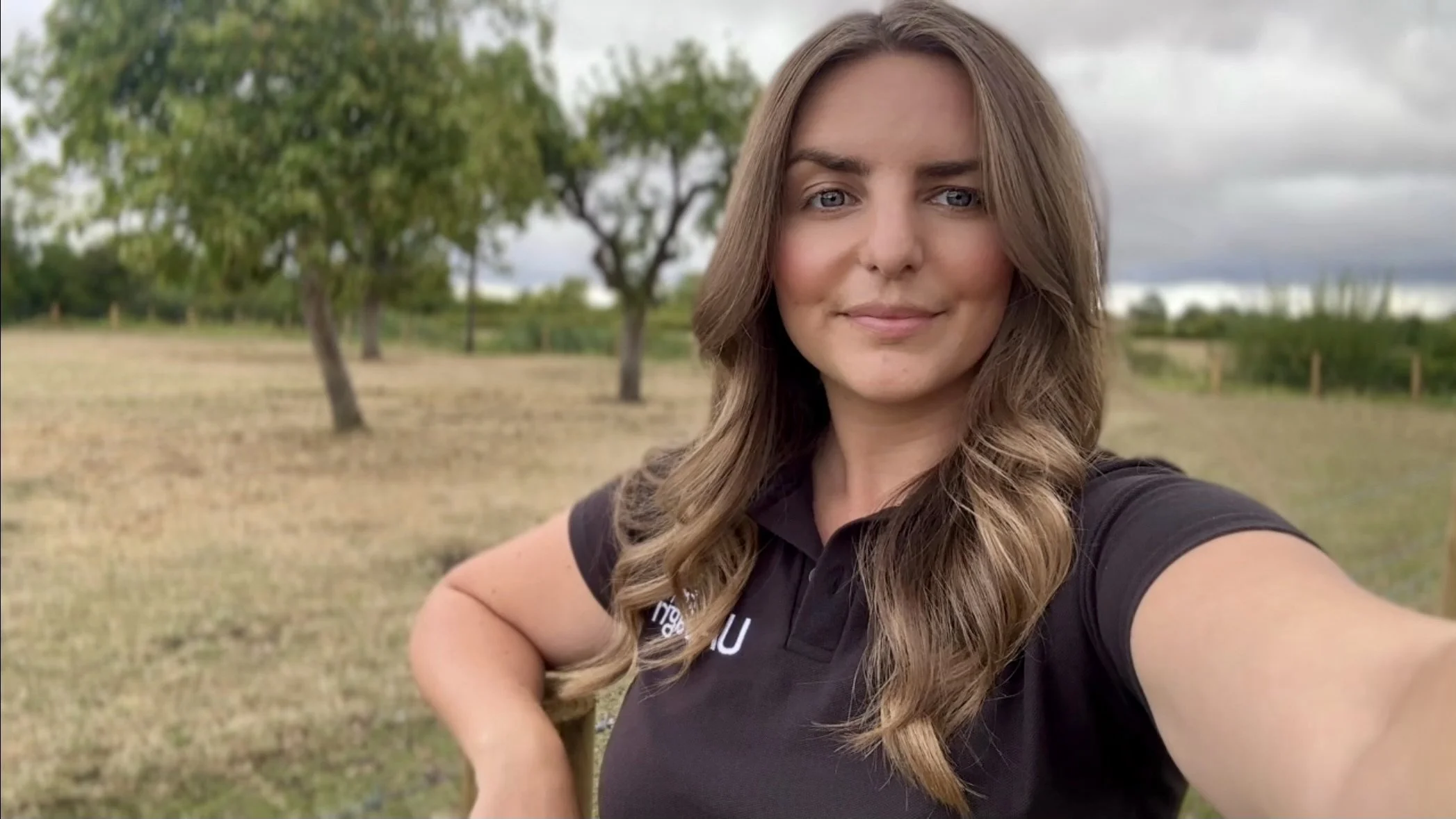 A woman with long wavy hair taking a selfie outdoors in a field with trees and cloudy sky in the background.