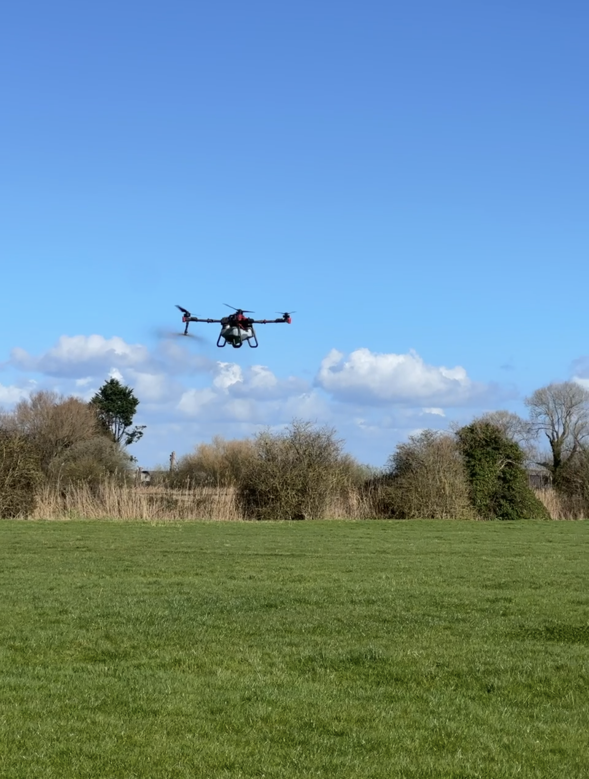 A quadcopter drone flying over a grassy field with trees and shrubs in the background under a blue sky with clouds.