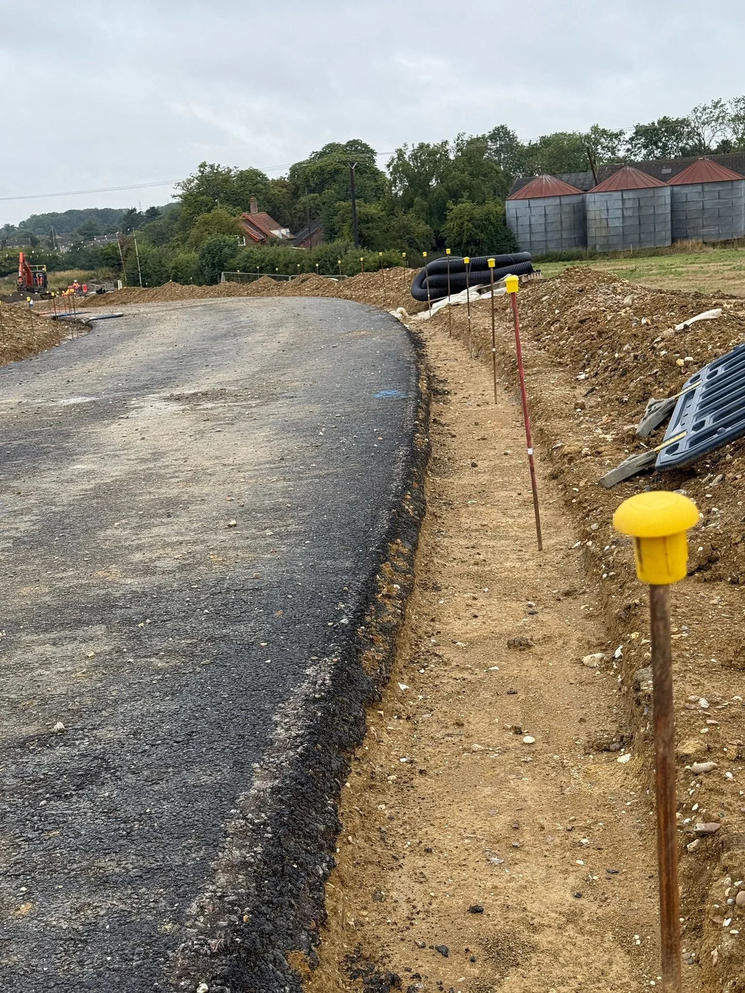 A dirt pathway runs alongside a paved road under construction, with orange and yellow marking pins and construction equipment visible in the background, amid a semi-rural landscape.