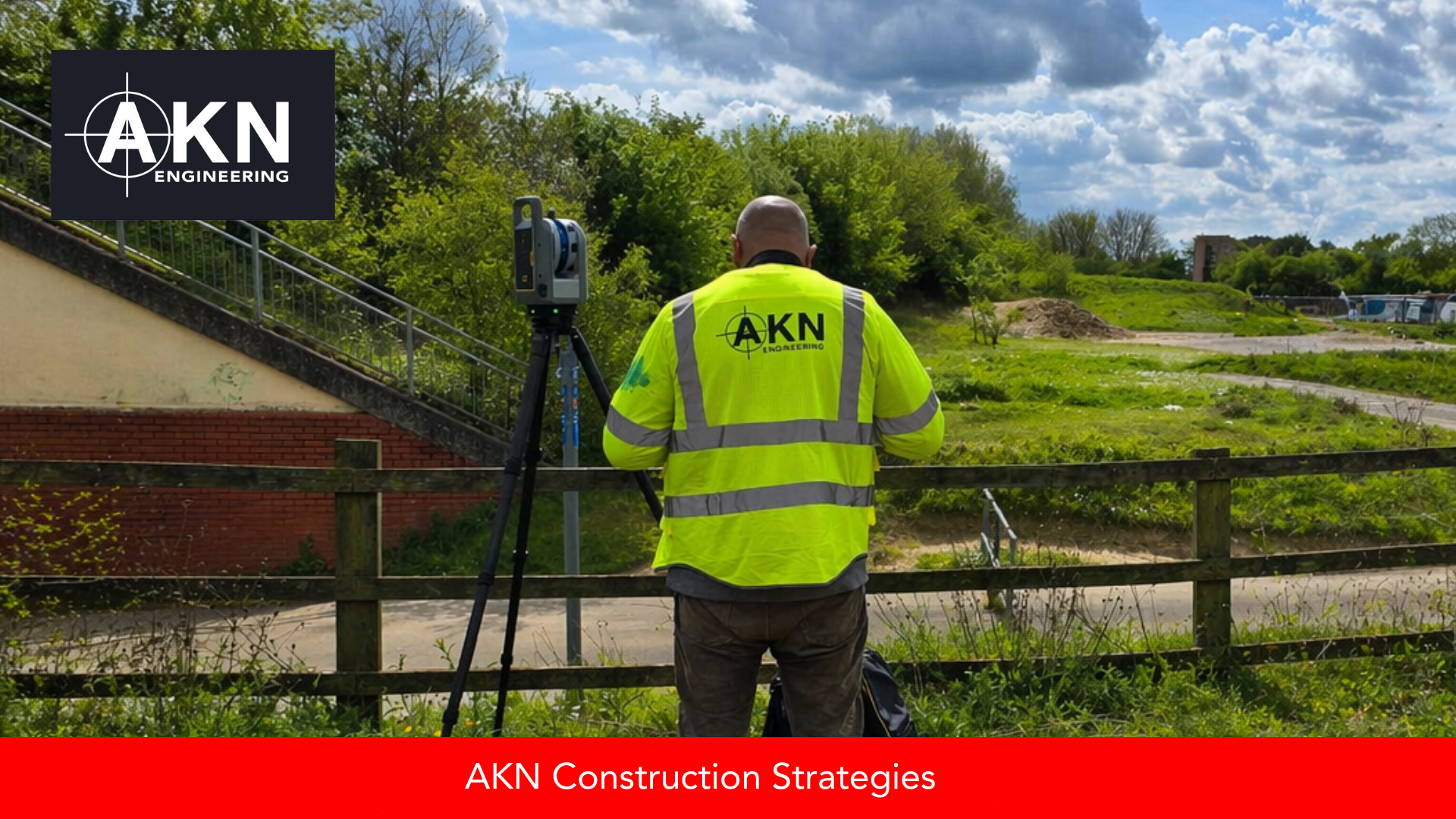 AKN Engineering operative in hi-vis PPE operating a Trimble laser scanner on tripod at a UK pre-construction site, with open ground and existing structures visible in the background.