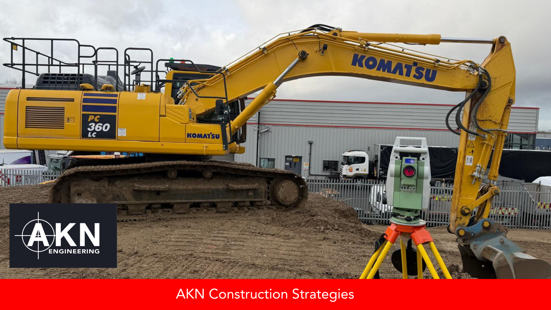 A total station on a yellow tripod set up on a levelled construction site alongside a tracked excavator, with AKN Engineering branding in the corner.
