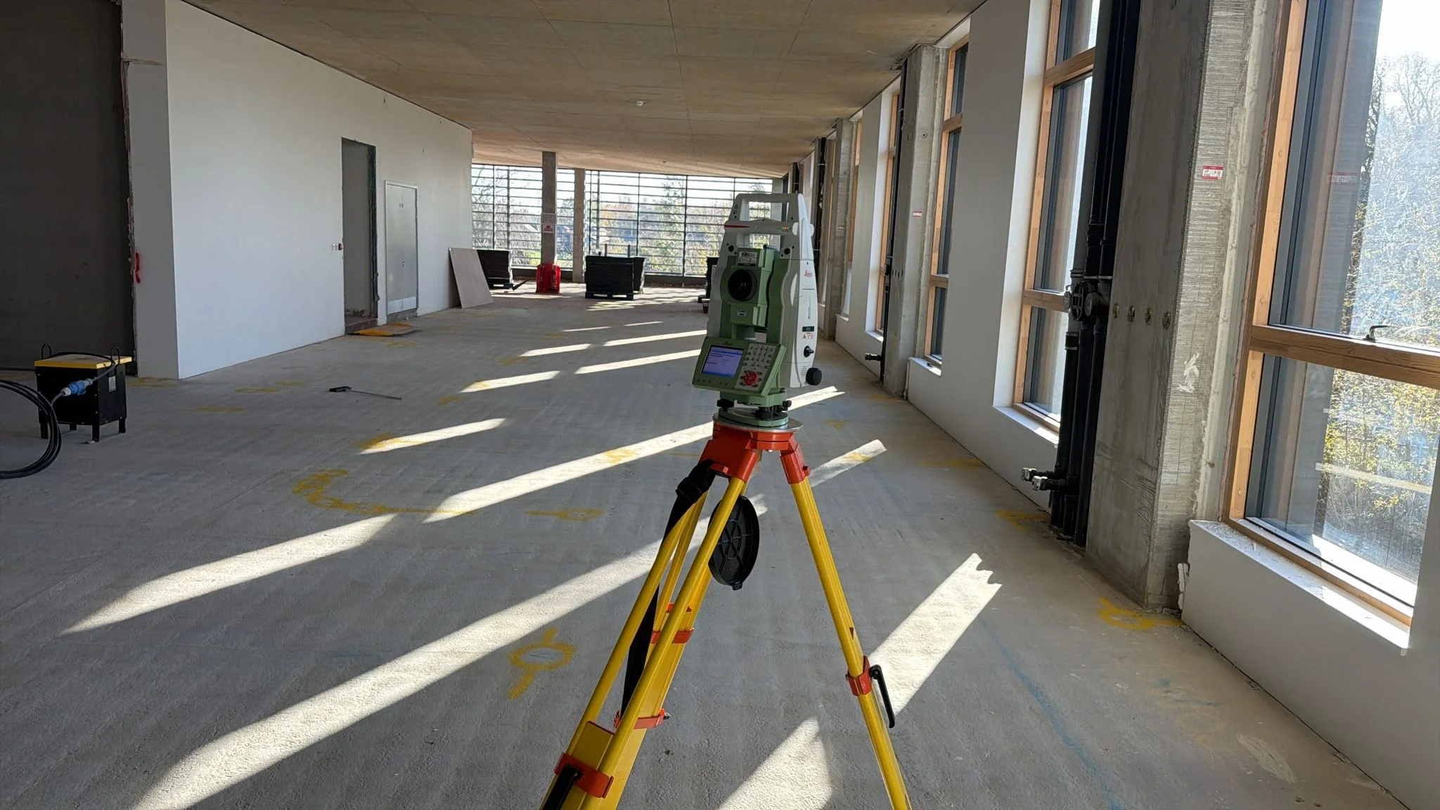 Interior of a building under construction with a surveyor instrument on tripod in the foreground, large windows on the right, and construction equipment and seating in the background.