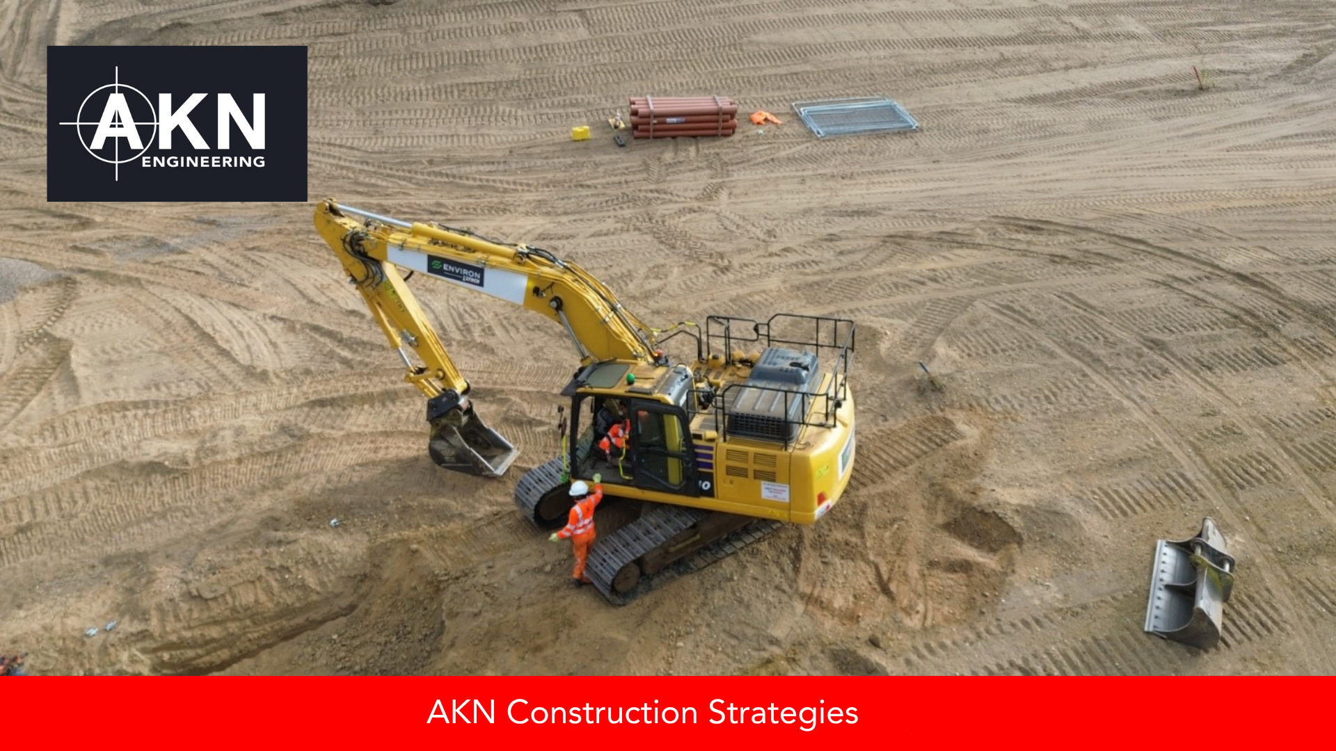 Aerial view of a UK construction groundworks site with a yellow excavator and operative in hi-vis PPE, piling auger visible in the foreground during early earthworks phase