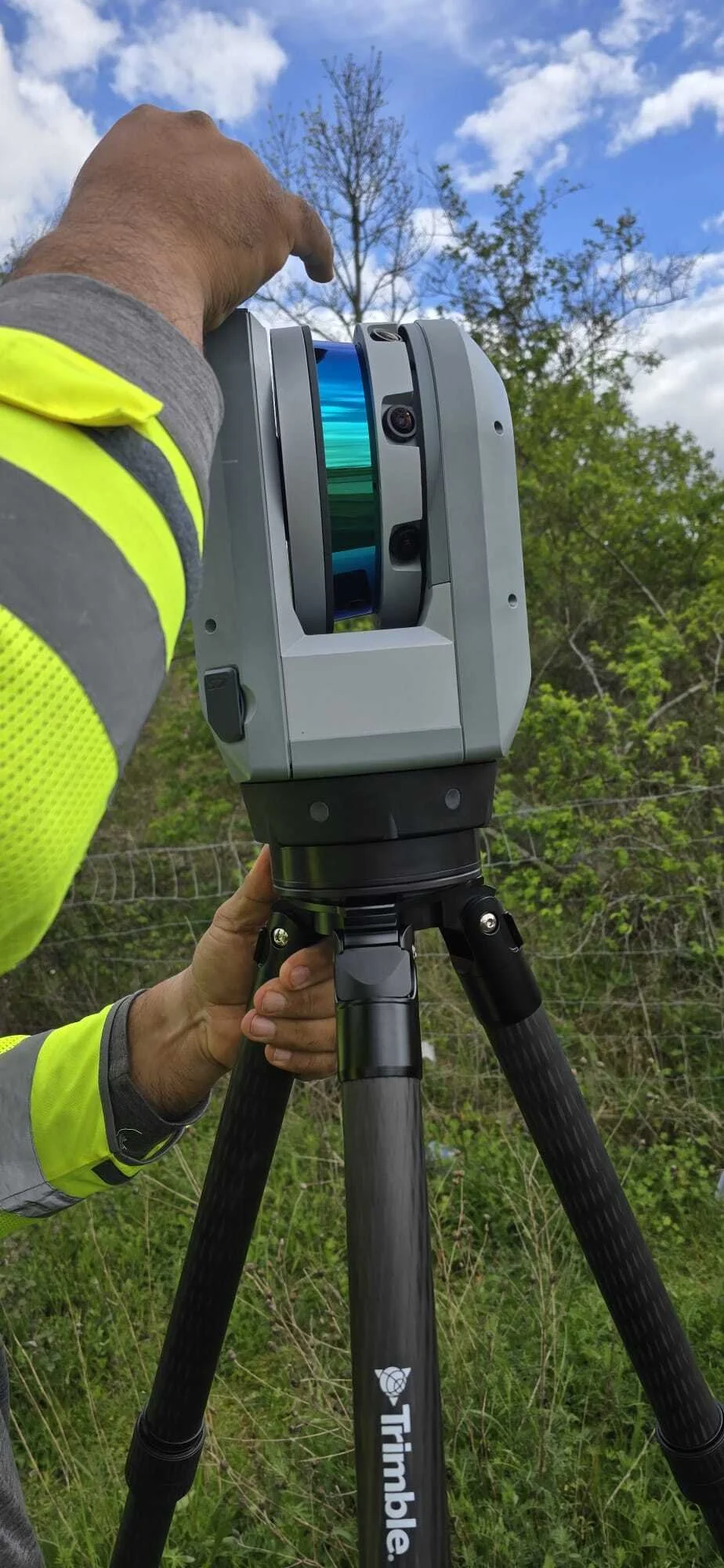 Trimble X9 3D laser scanner being operated in a field, you can see the operators arms, wearing yellow PPE
