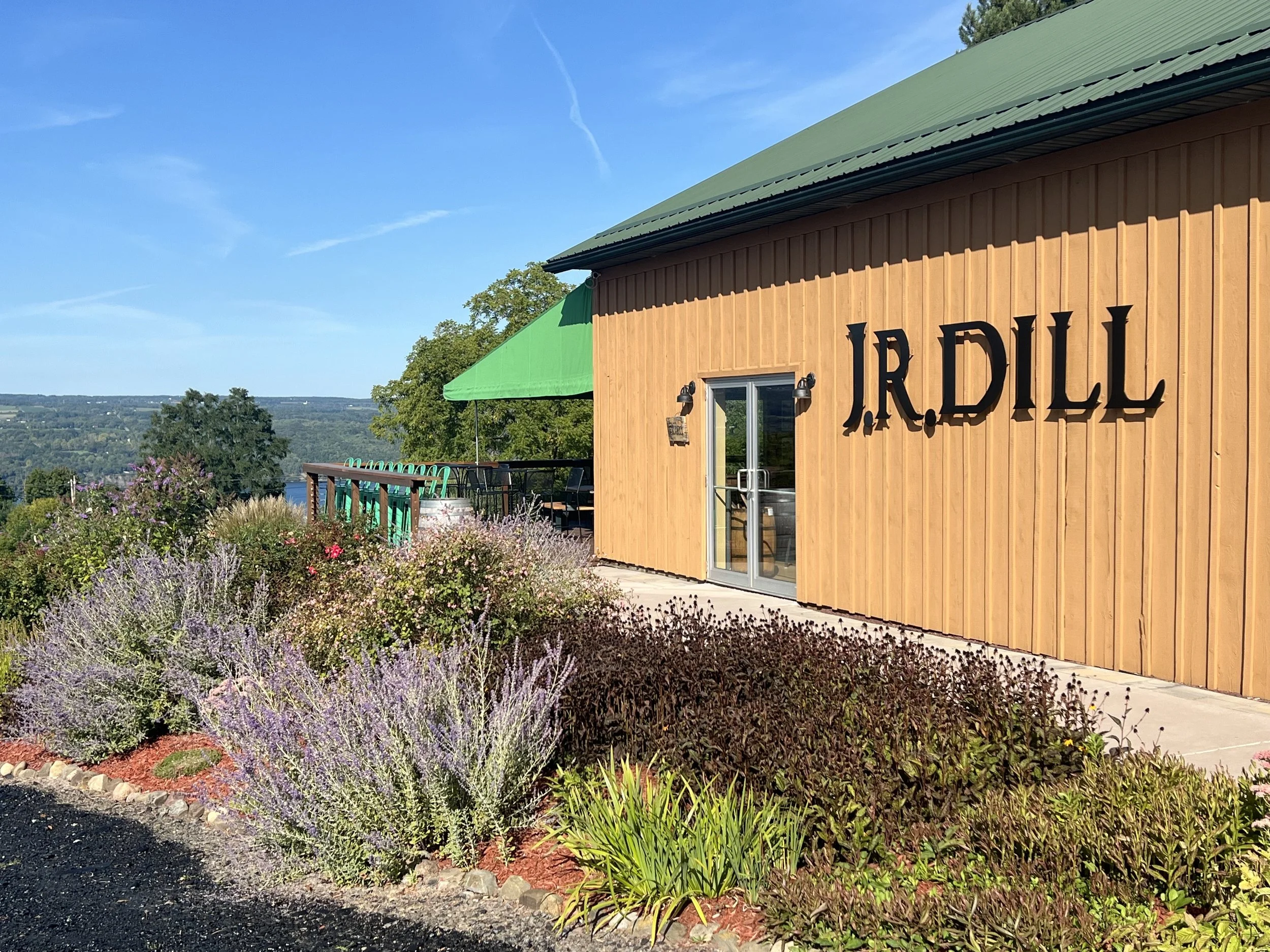 View of a building with the sign 'J.R. DILL' with a landscaped garden, a deck with seating and a green canopy, overlooking a scenic landscape.