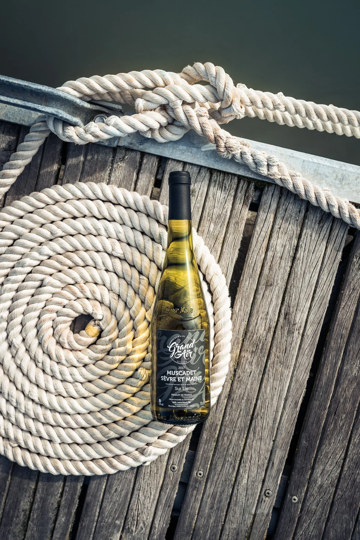 Une bouteille de vin blanc posé sur un matelas de corde enroulée, avec un pont en bois et de l'eau en arrière-plan.