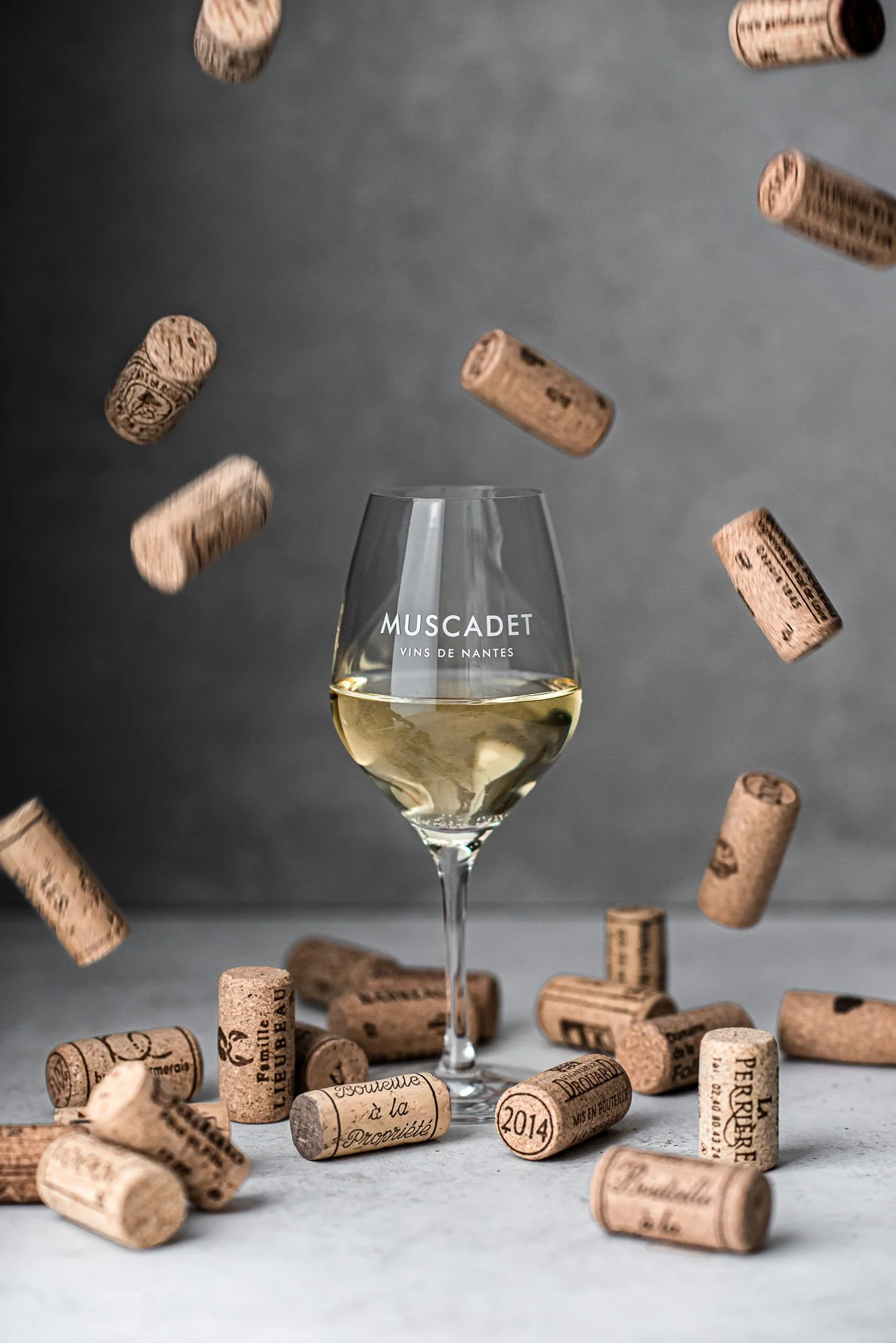 Verre de vin blanc avec le mot 'MUSCADET' et 'VINS DE NANTES' écrit dessus, entouré de bouchons de vin en liège en suspension et sur la surface, avec un fond gris.