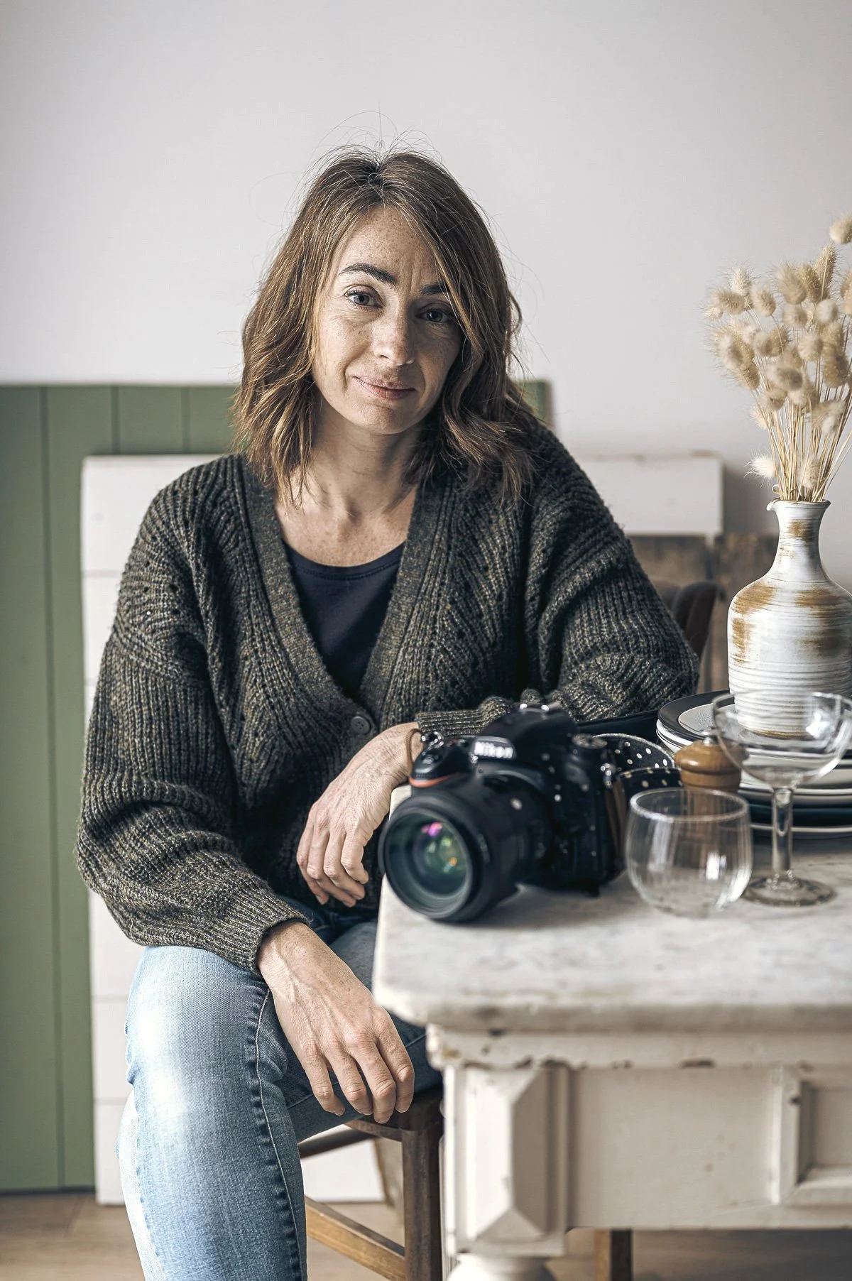 Femme assise à une table avec un appareil photo, vaisselle et vase avec fleurs en arrière-plan.
