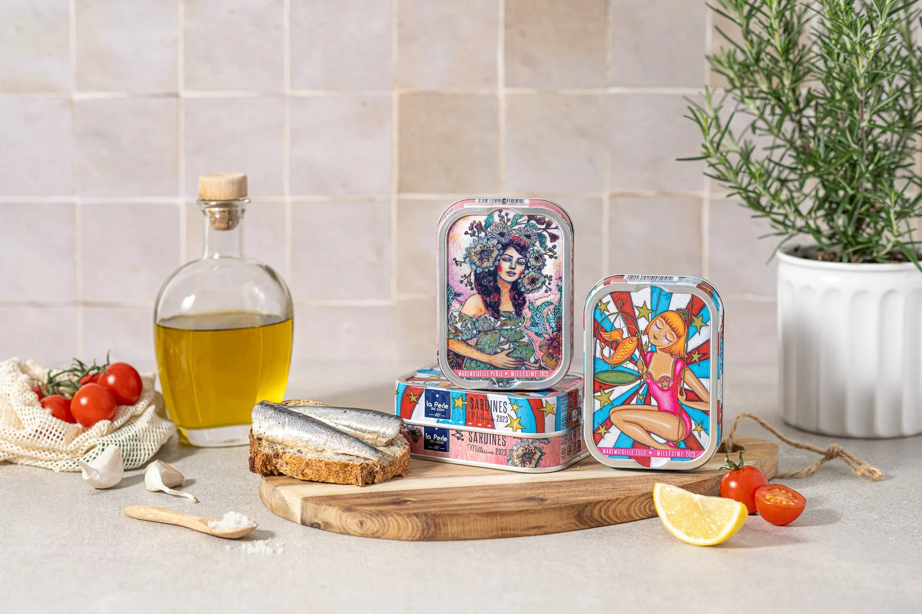 Boîtes de conserve illustrées avec des dessins de femmes, une bouteille d'huile d'olive, tomates cerises, citron en quartier, pain garnie d'anchois, une branche de thym ou romarin dans un pot blanc, et une tomate cerise, sur un plan de travail en cuisine avec une ardoise en bois.