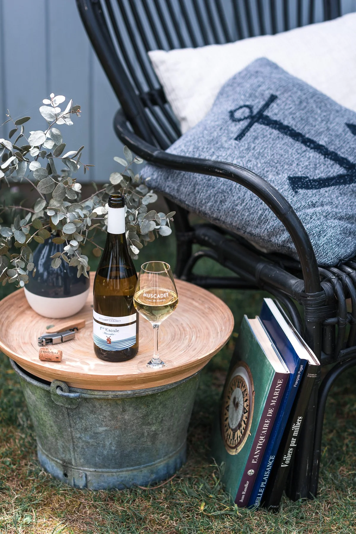 Une balancelle noire avec un coussin gris et une couverture avec une ancre, un tabouret en métal avec une planche en bois dessus, un livre de marine, une bouteille de vin blanc, un verre de vin blanc, un vase avec des feuilles vert gris, des clés et 