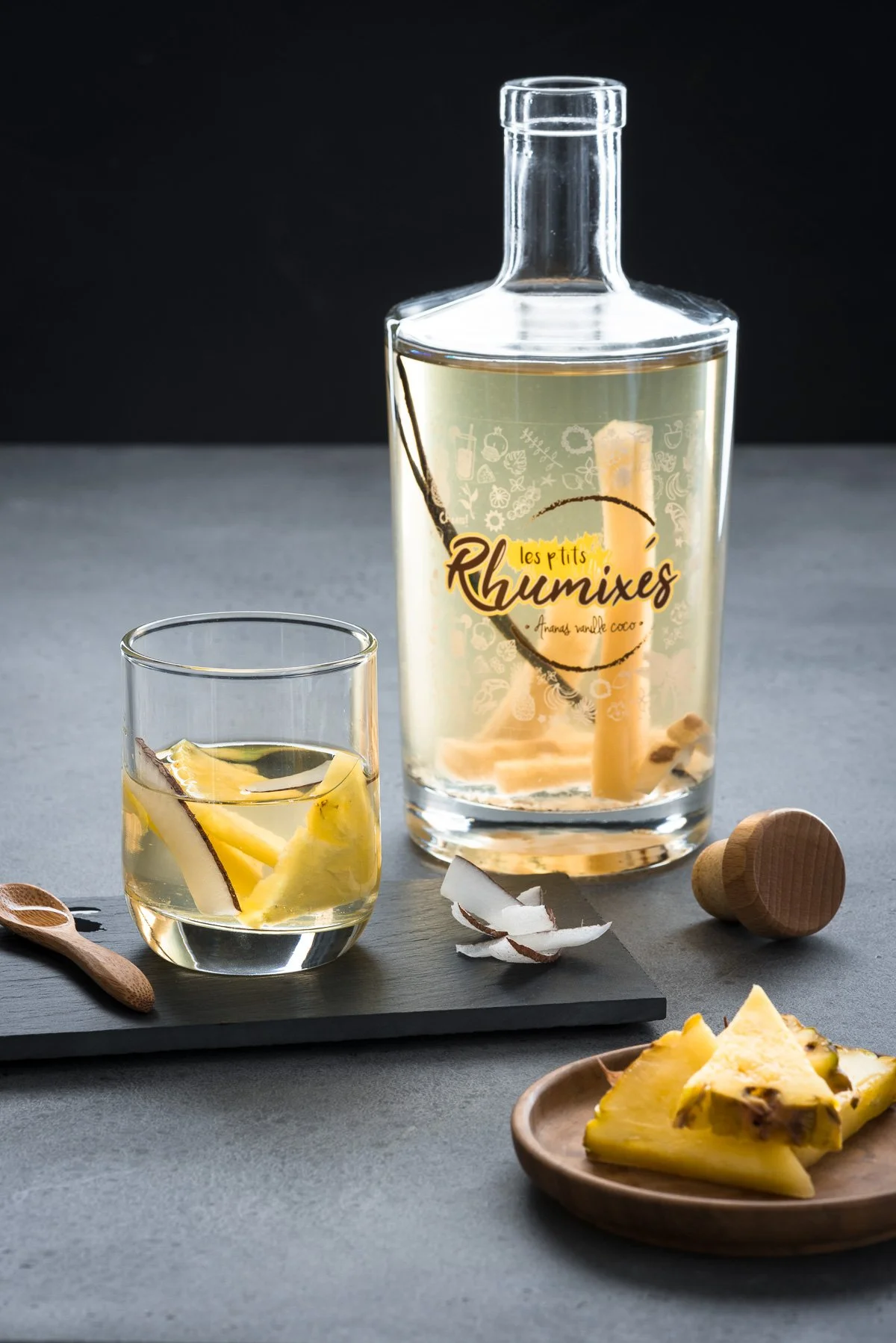 Une bouteille de rhum arrangé accompagnée d'un verre rempli de rhum avec des morceaux d'ananas et de noix de coco, posé sur une planche en bois, avec des morceaux de noix de coco et un morceau d'ananas sur une petite assiette en bois.