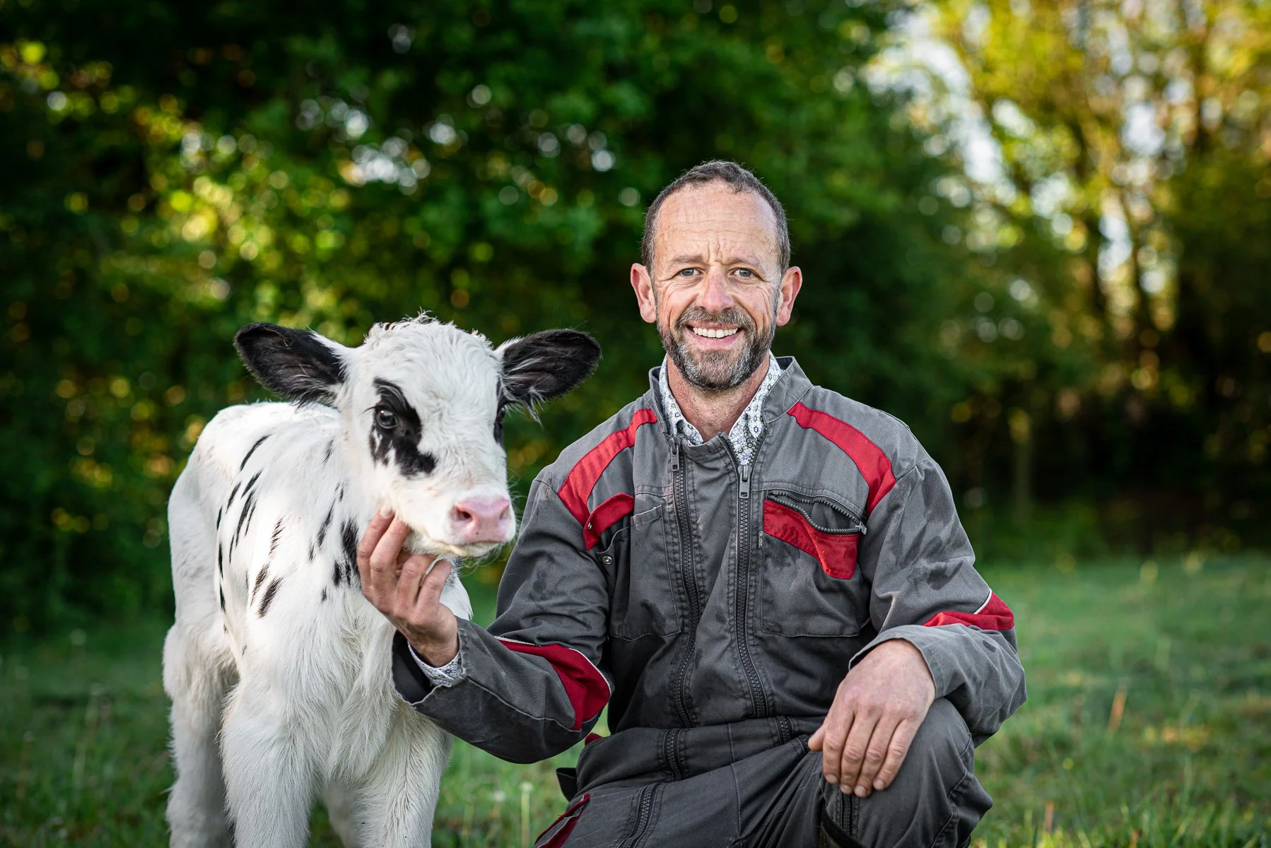 Un homme souriant en combinaison de travail, tenant la tête d'une jeune vache noire et blanche dans un pré verdoyant, avec un fond de arbres verts.