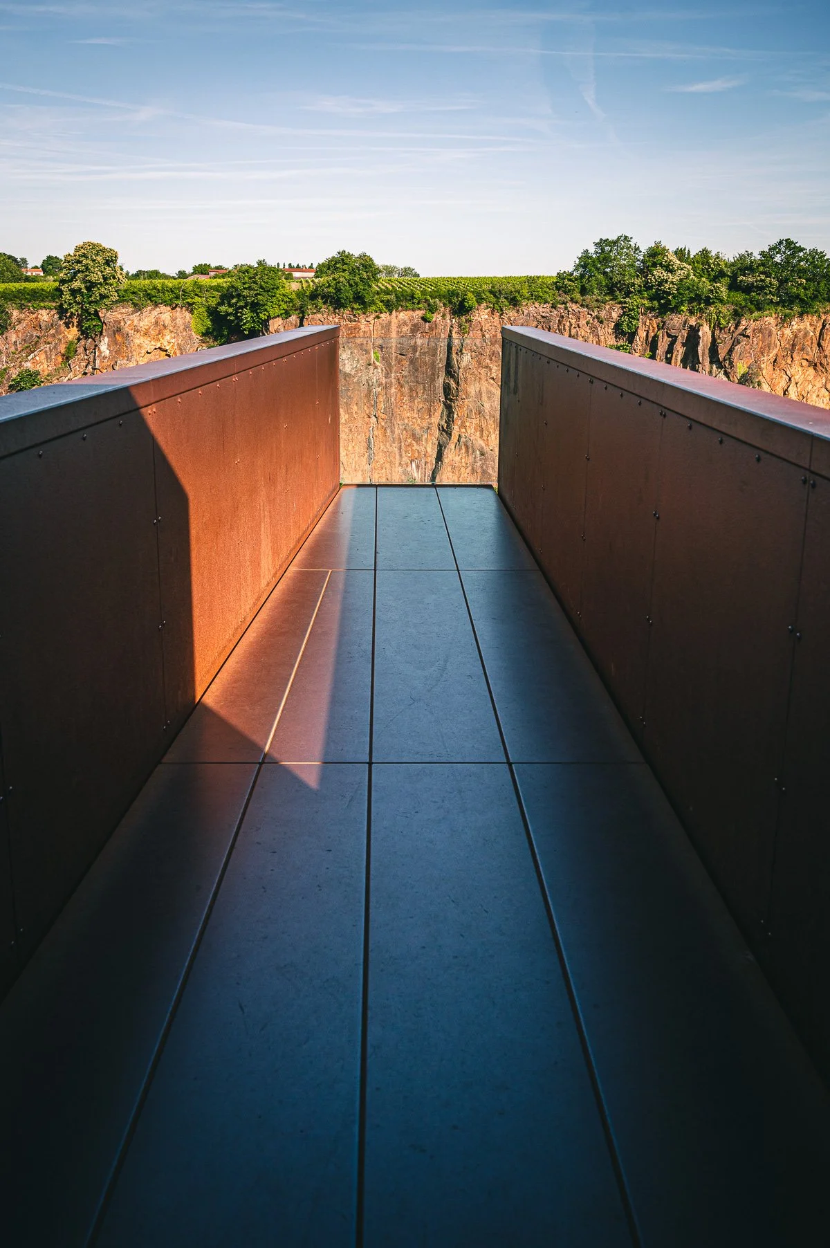 Une passerelle en métal donnant sur une falaise rocheuse avec un panorama de arbres et un ciel bleu clair.
