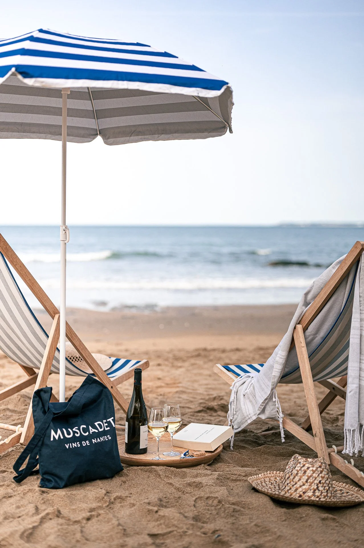 Plage avec deux chaises longues, un parasol, un sac, une bouteille de vin, deux verres, un livre, un chapeau de paille, et l'océan en arrière-plan.